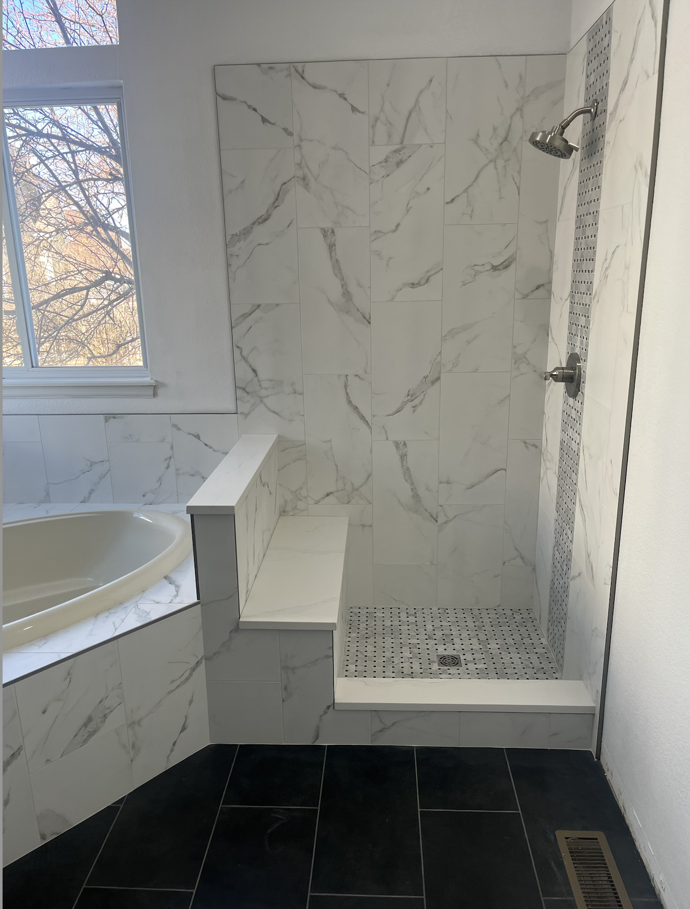 A modern bathroom with a marble shower area, a bathtub with marble sides, a window showing trees outside, and black tiled flooring.