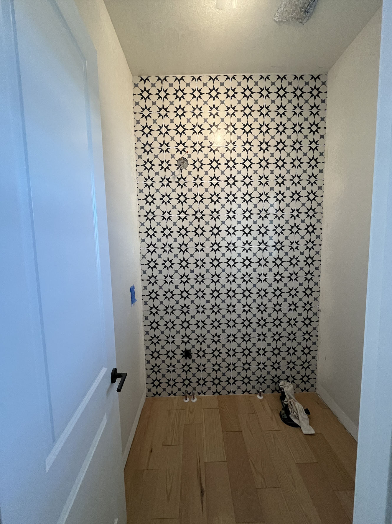 A room with a patterned black and white tile accent wall on the back, a light-colored hardwood floor, and a white door on the left side. Tools and supplies for installation or renovation are visible on the floor near the wall.