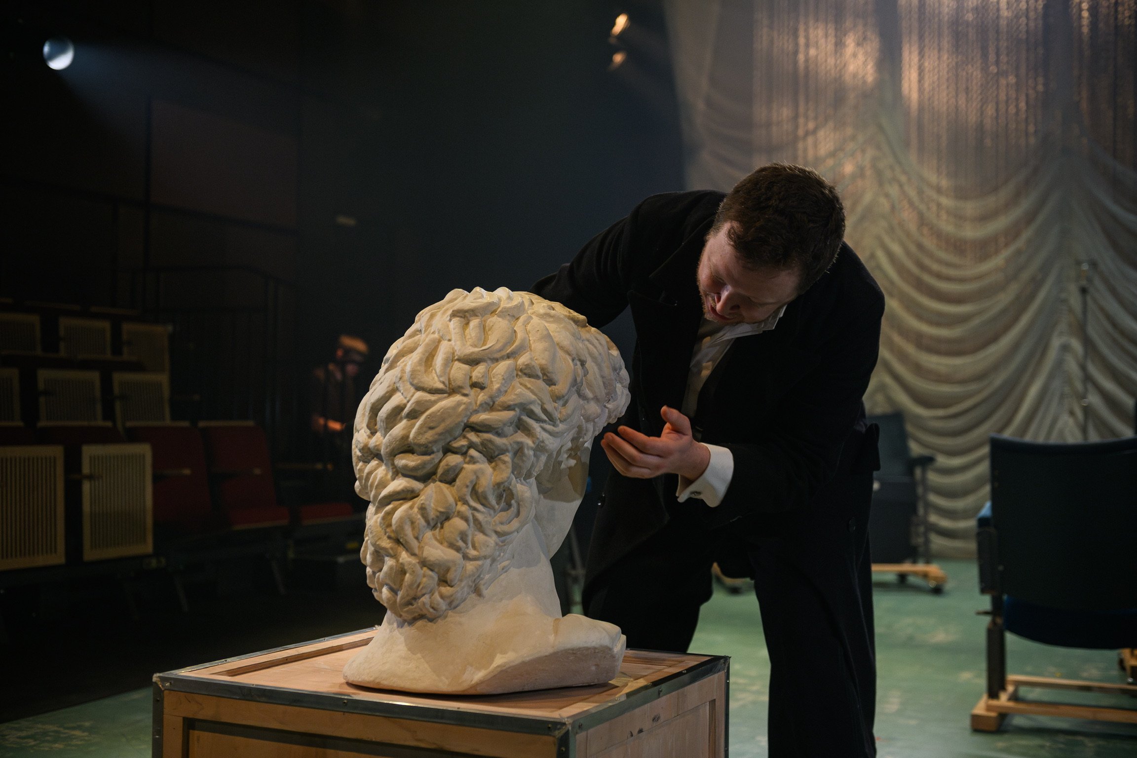 A man in formal attire closely examines a large, detailed sculpture of a human brain in an art gallery or theater setting with red theater seats and ornate curtains in the background.