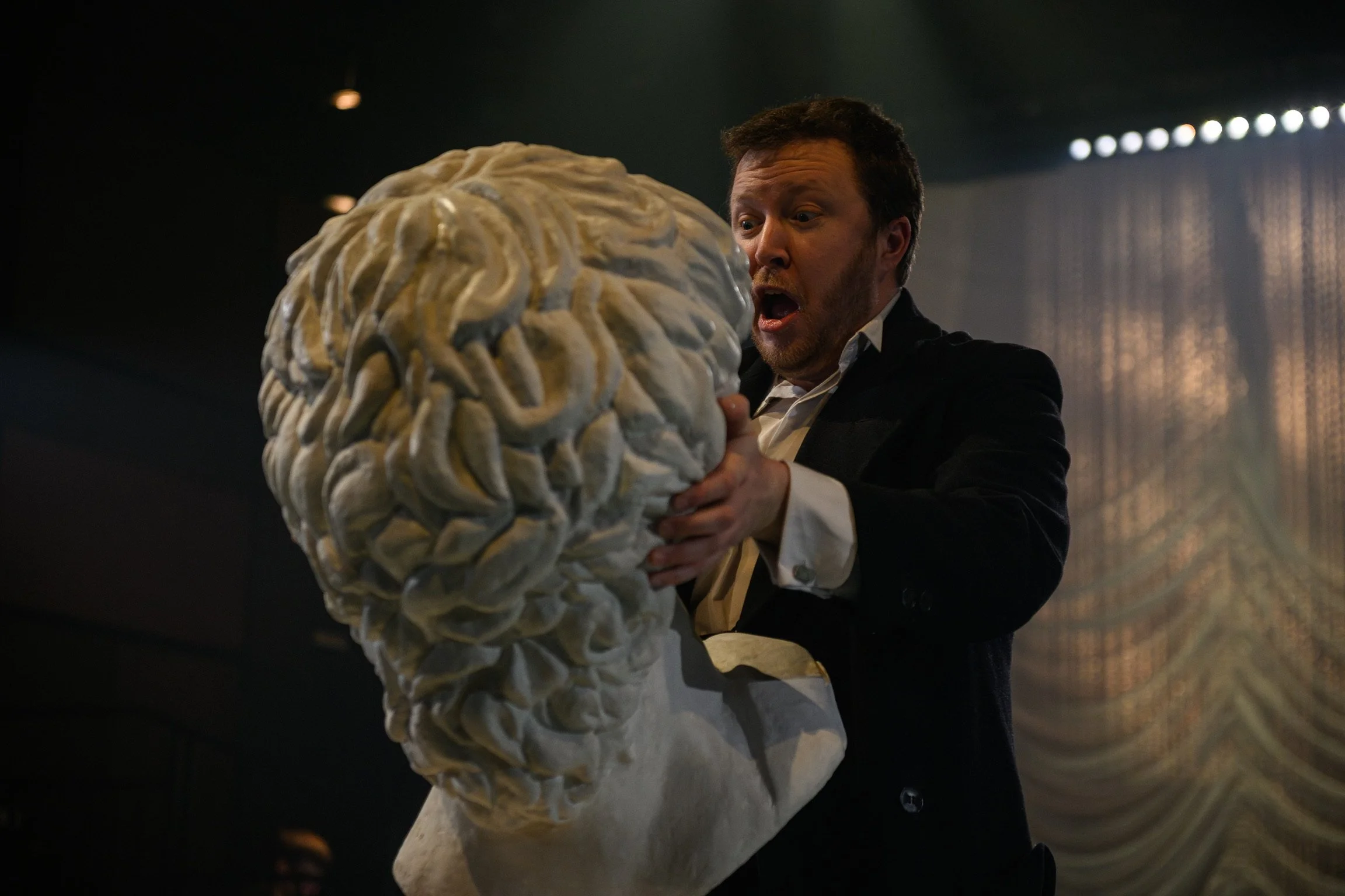 A man with a beard and dark hair, dressed in formal attire, holds a large, detailed model of a human brain as he exhibits surprise or excitement in a dimly lit room.