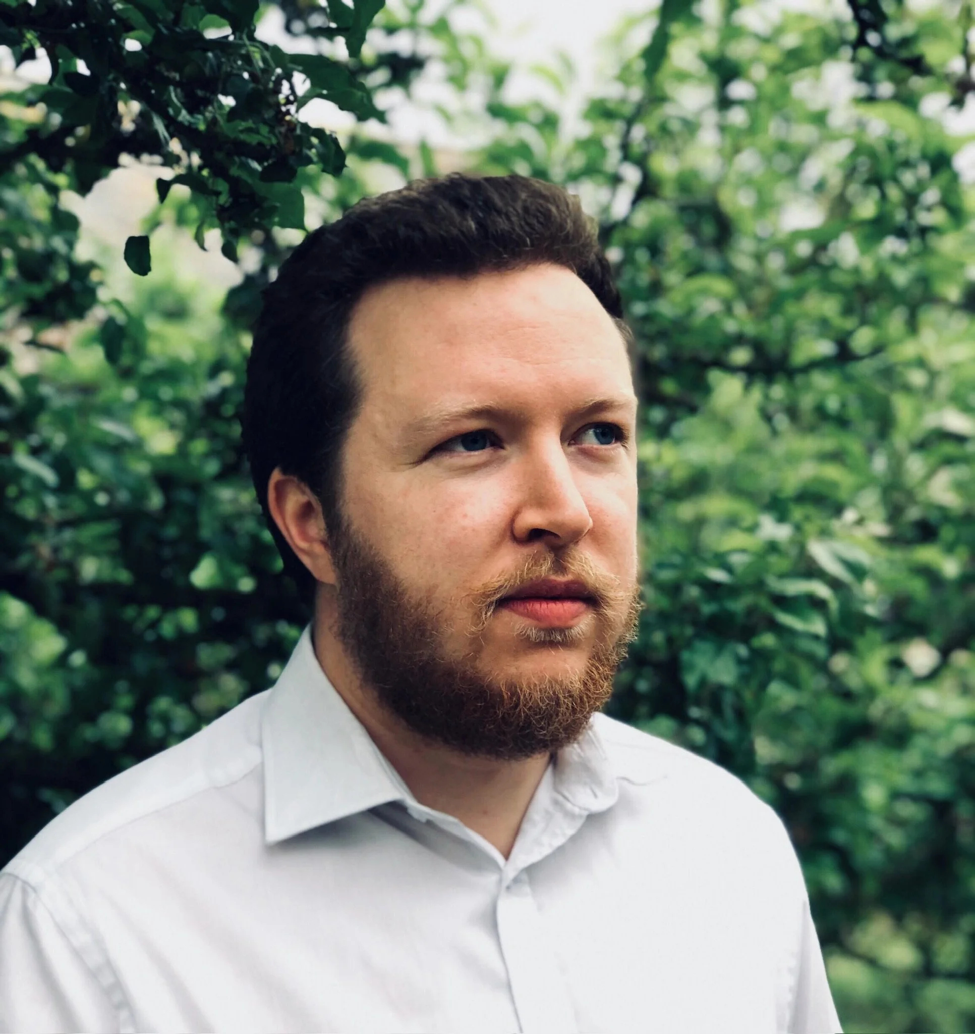 Close-up of a man's face with dark hair and beard, wearing a white collared shirt, outdoors in a green leafy environment.