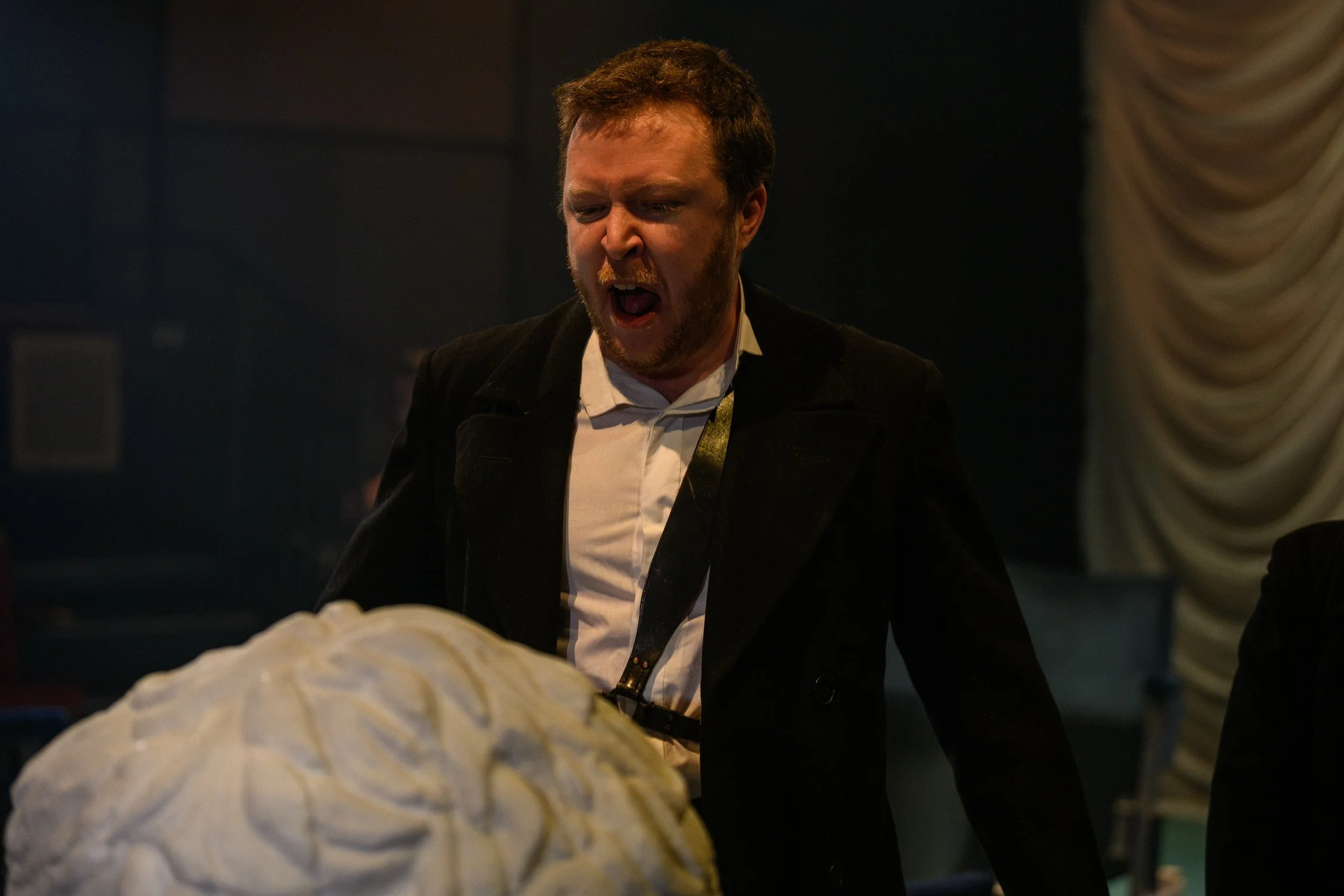 Man with ginger hair and beard, dressed in a black jacket and white shirt, appears to be shouting or singing passionately. He is in a dark room with a dark background and some draped fabric on the right side.