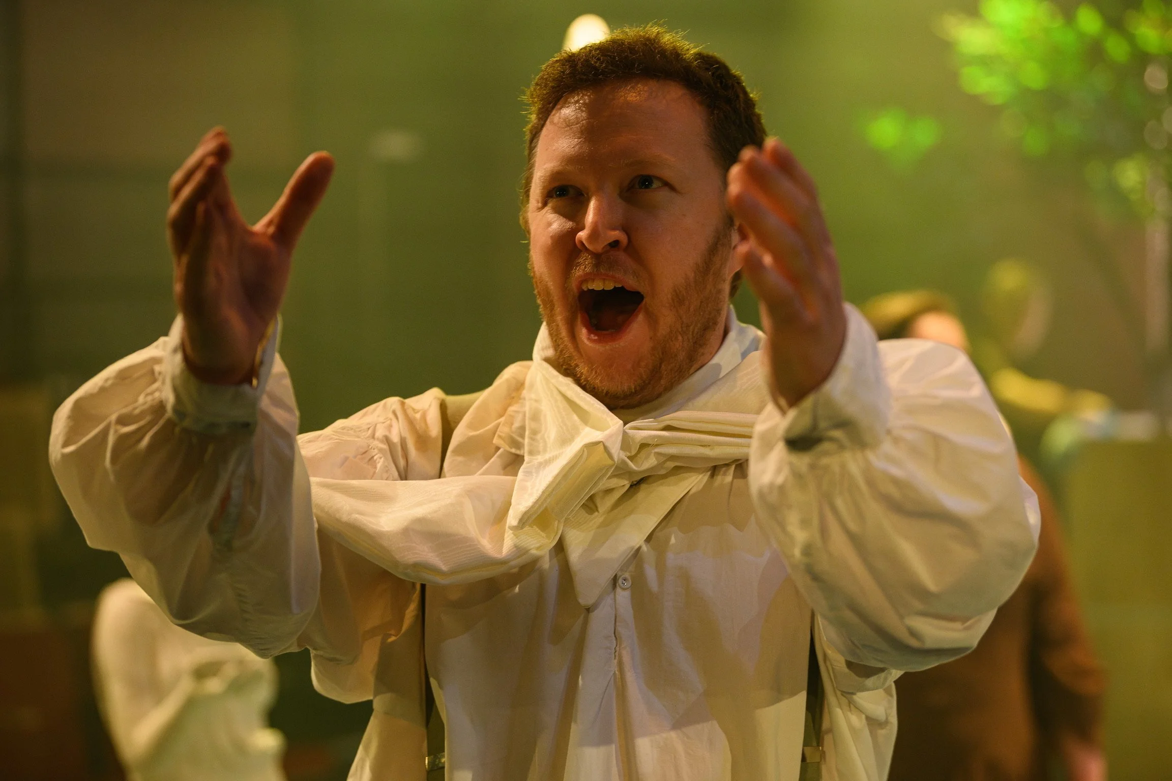A man with short brown hair and facial hair, wearing a cream-colored shirt, gestures with his hands and has his mouth open as if speaking or shouting. The background is blurred with green and yellow tones.