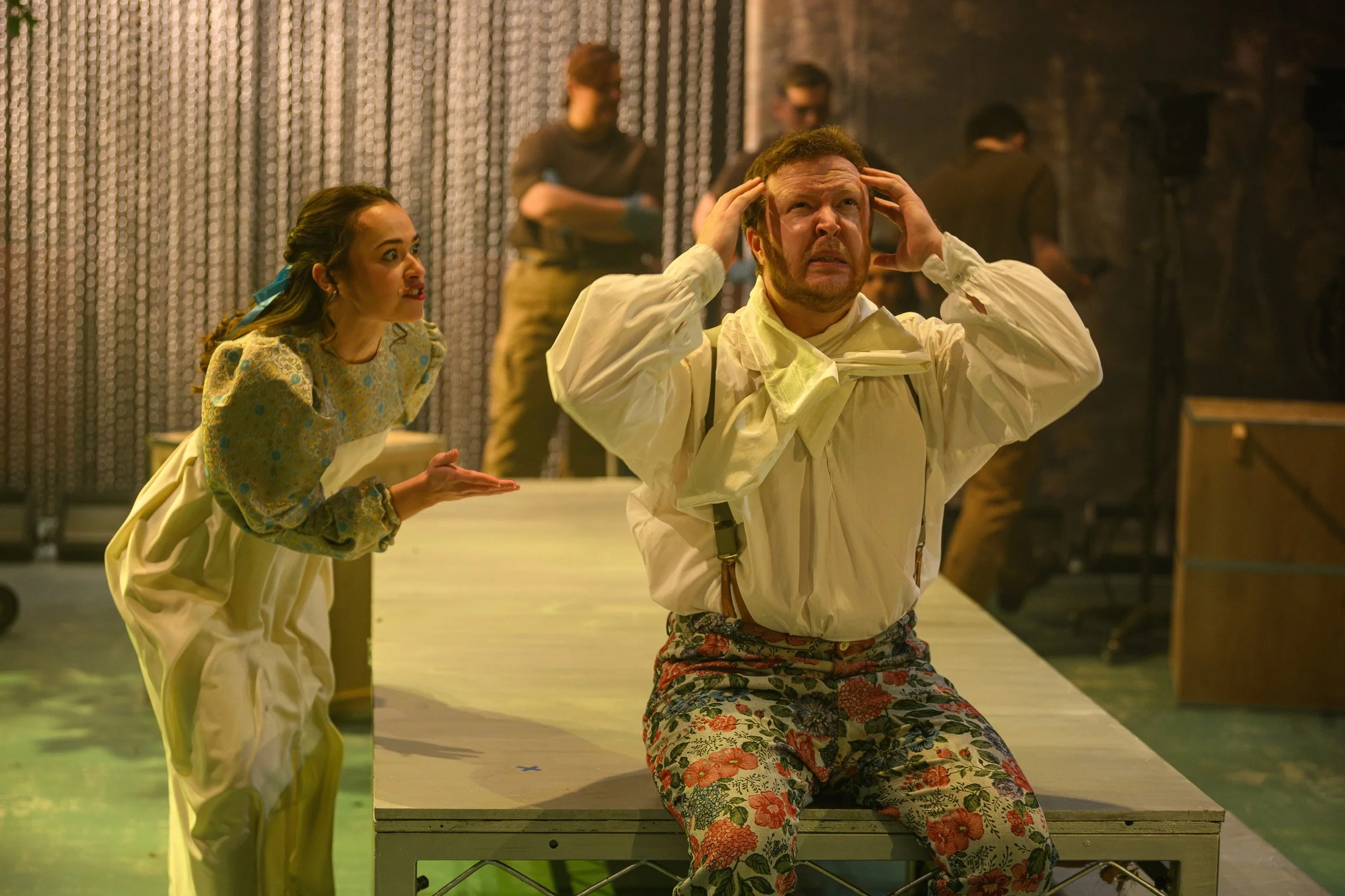 A man appears distressed while seated at a table with his hands on his head. Next to him, a woman with a concerned expression gestures towards him. The background features a metallic curtain and multiple people, suggesting a scene from a theatrical p