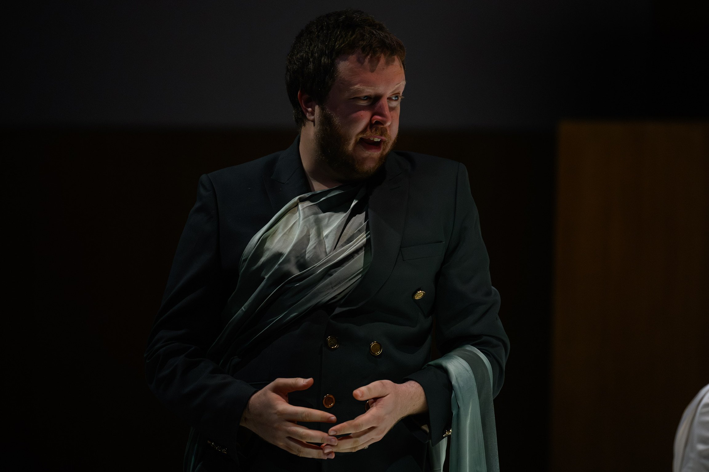 A man in a dark suit with a feathered sash stands in a dark room, speaking or reacting to something, with a serious expression on his face.