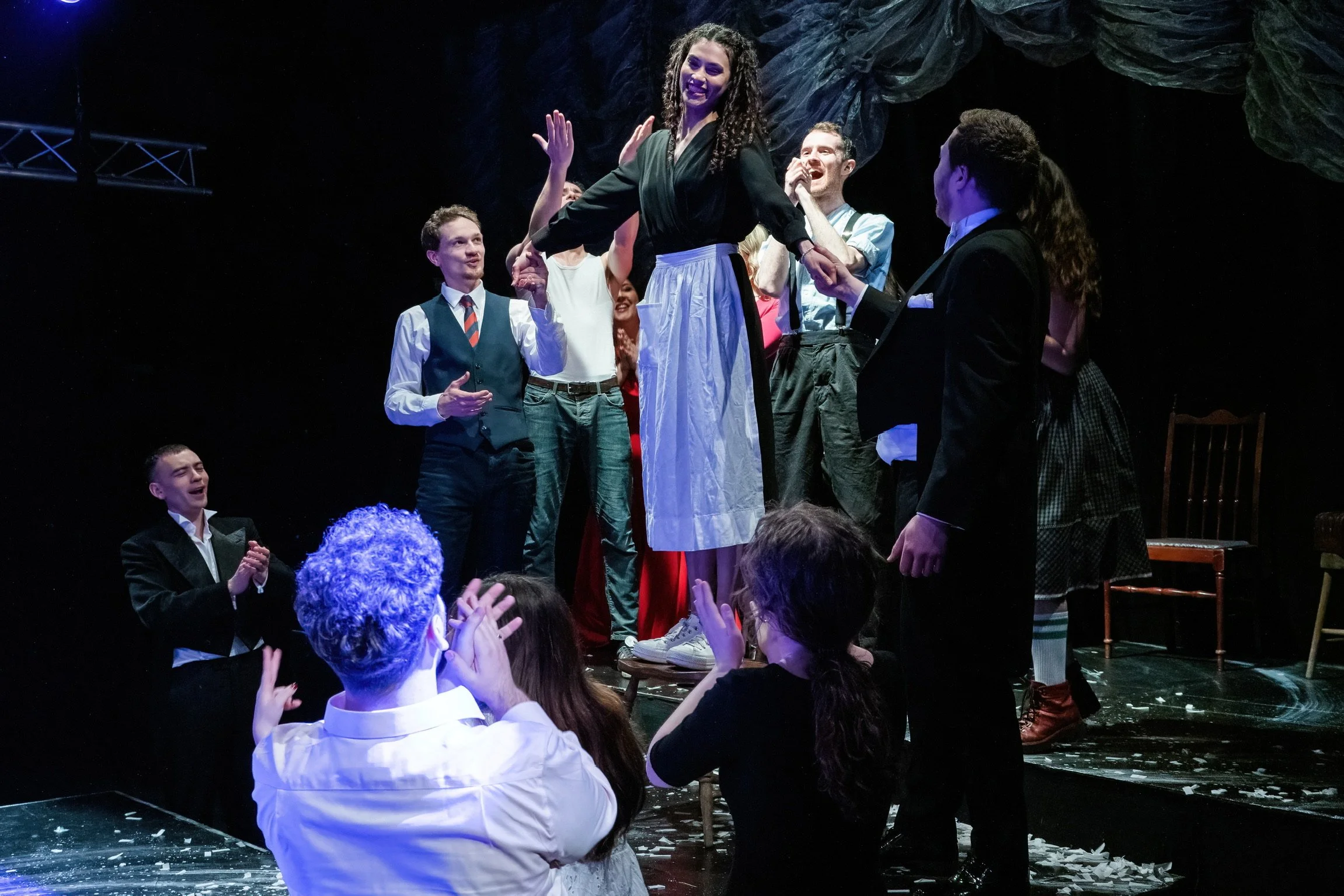 Stage scene with a woman on a raised platform surrounded by people clapping and smiling, with theatrical lighting and dark background.
