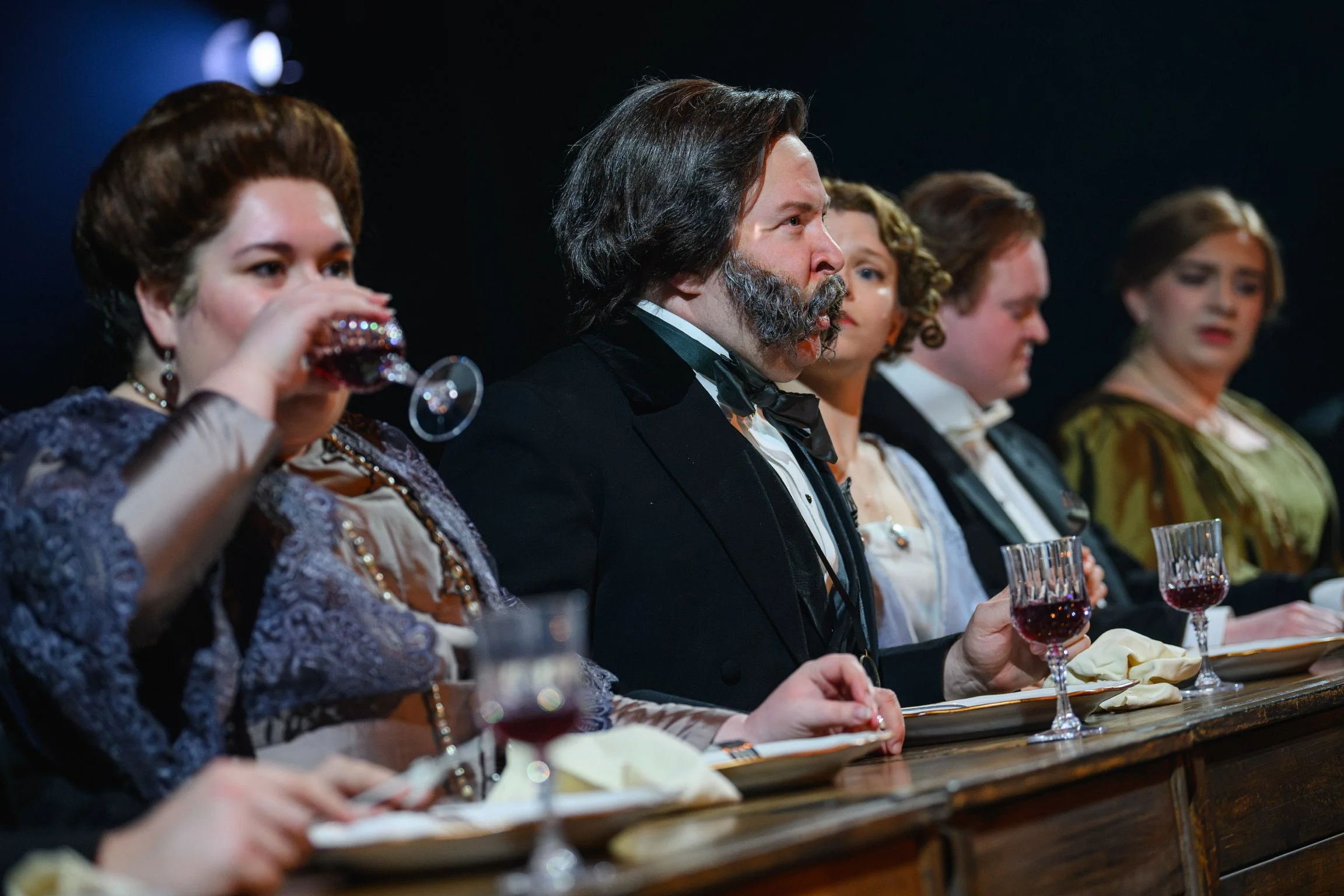 People dressed in vintage formal attire sitting at a long table with glasses of red wine, at a formal event or dinner.