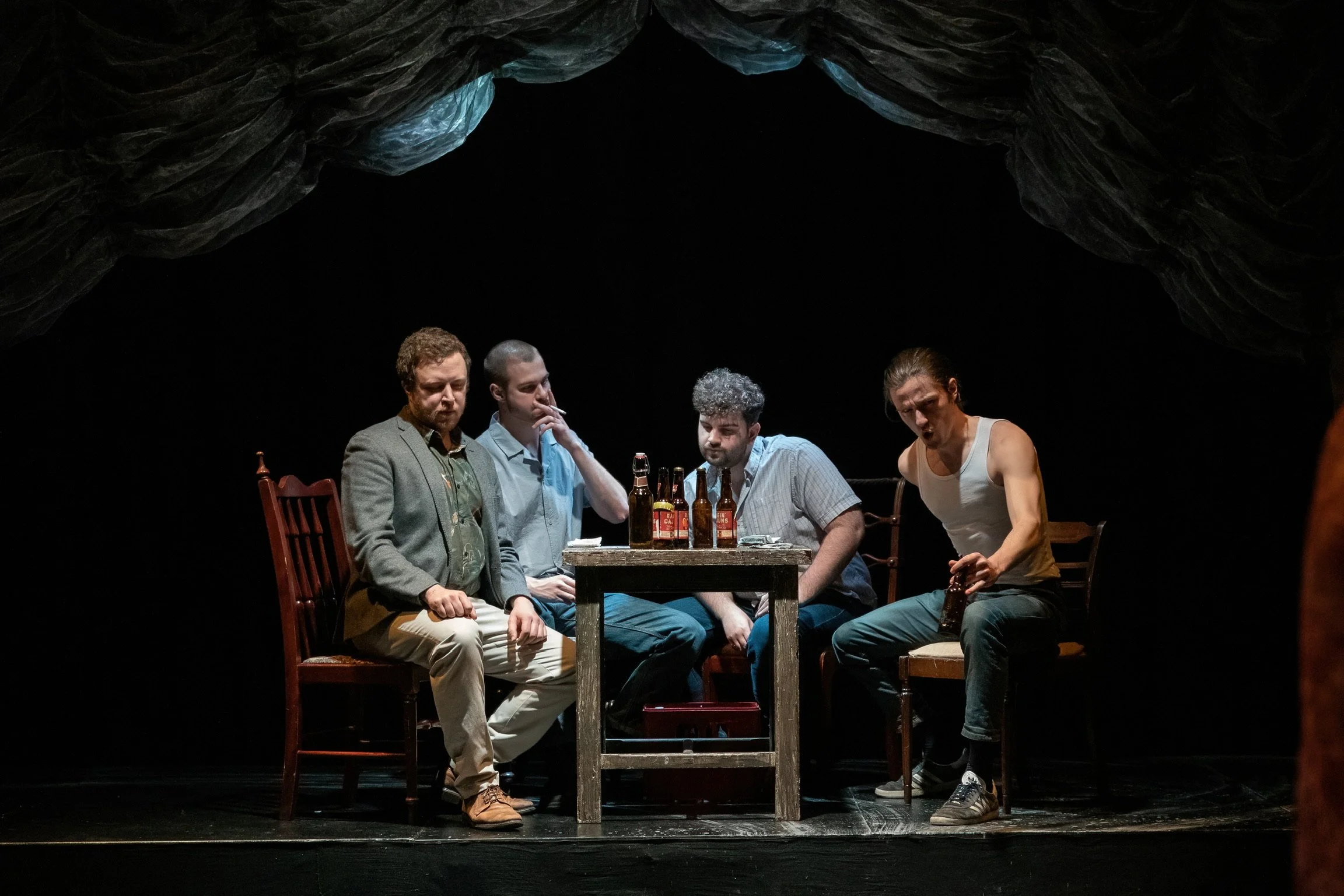 Four men sitting around a table on a stage with a dark background and draped fabric above them. One man is wearing a blazer, and the others are dressed casually. The table has several beer bottles, and one man is leaning forward with an animated expr