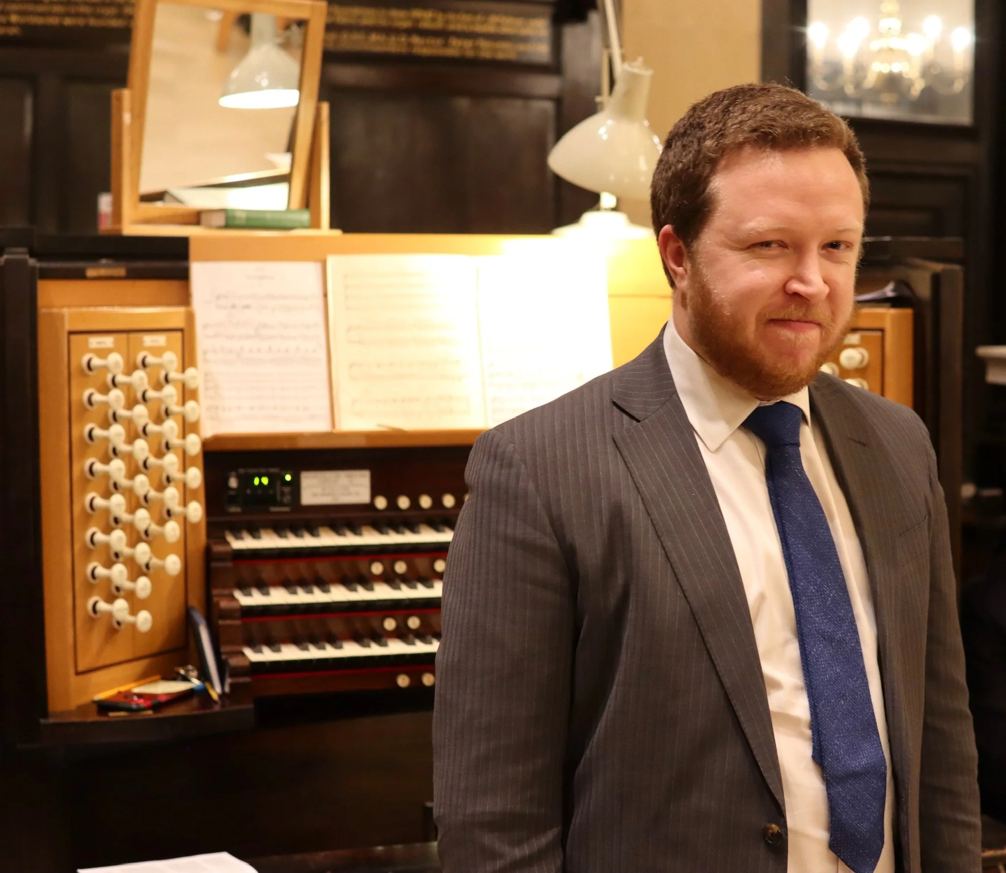 A man in a business suit with a bow tie standing in front of an organ or a pipe organ in a formal setting.