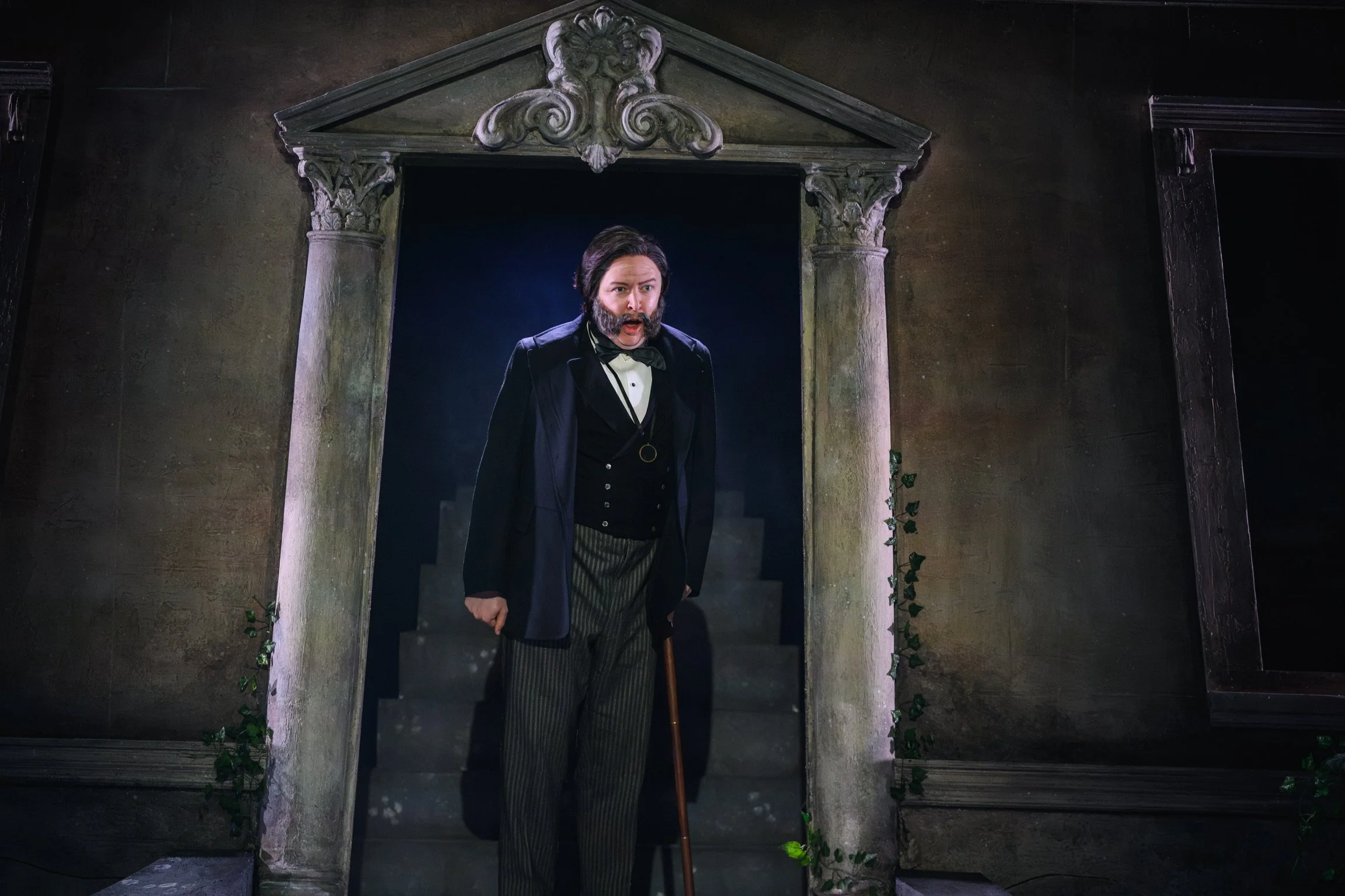 A man dressed as a character from a historical or theatrical setting, with long hair, a beard, wearing a tuxedo with a bow tie, standing in a doorway with ornate stone columns and a triangular pediment above, holding a cane or walking stick.
