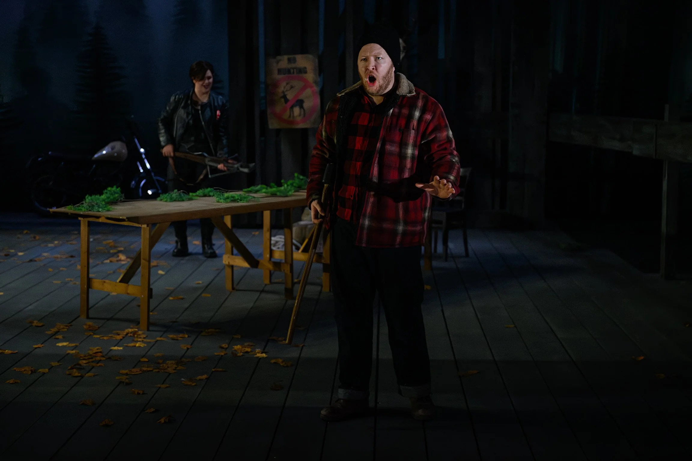 A man with a beard, wearing a black beanie and red plaid jacket, standing on a wooden deck at night, yelling, with a woman in a black leather jacket in the background, and a table with green plants and fallen leaves around.