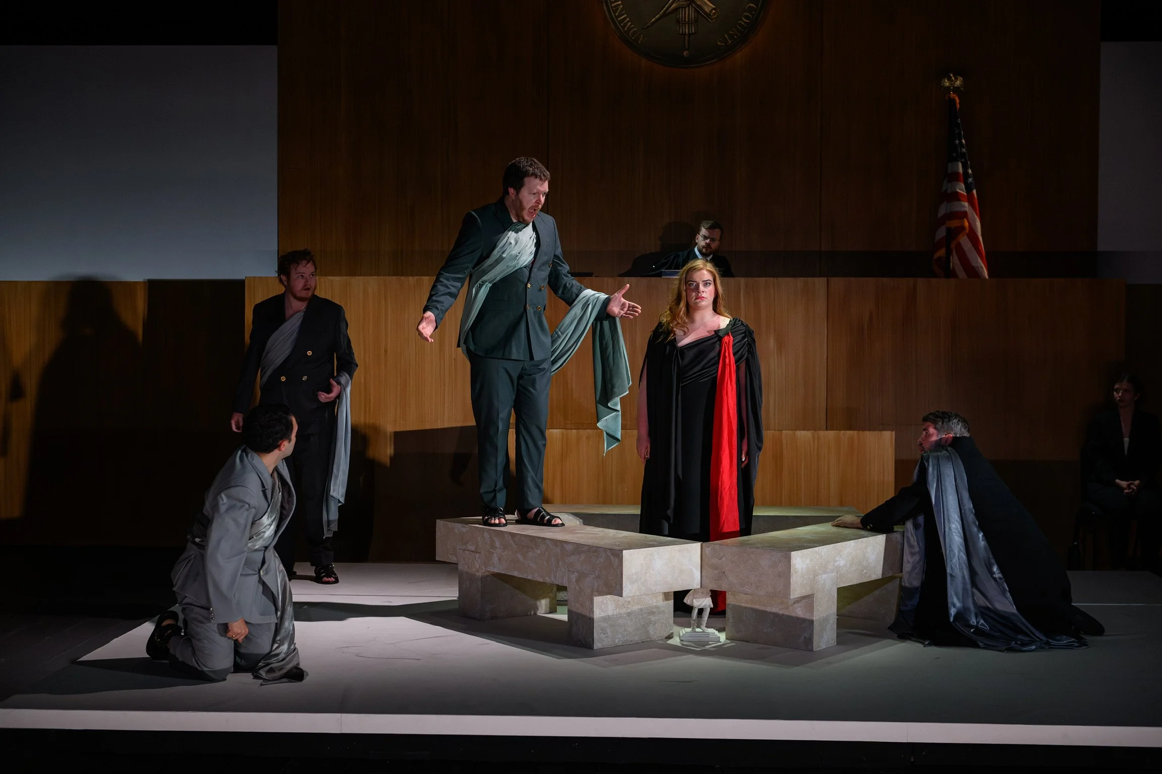 A theatrical performance depicting a courtroom scene with actors in costume. A man in a dark suit with a light-colored scarf gestures dramatically while standing on a raised platform. A woman in a black gown with a red sash looks solemnly. Several ot