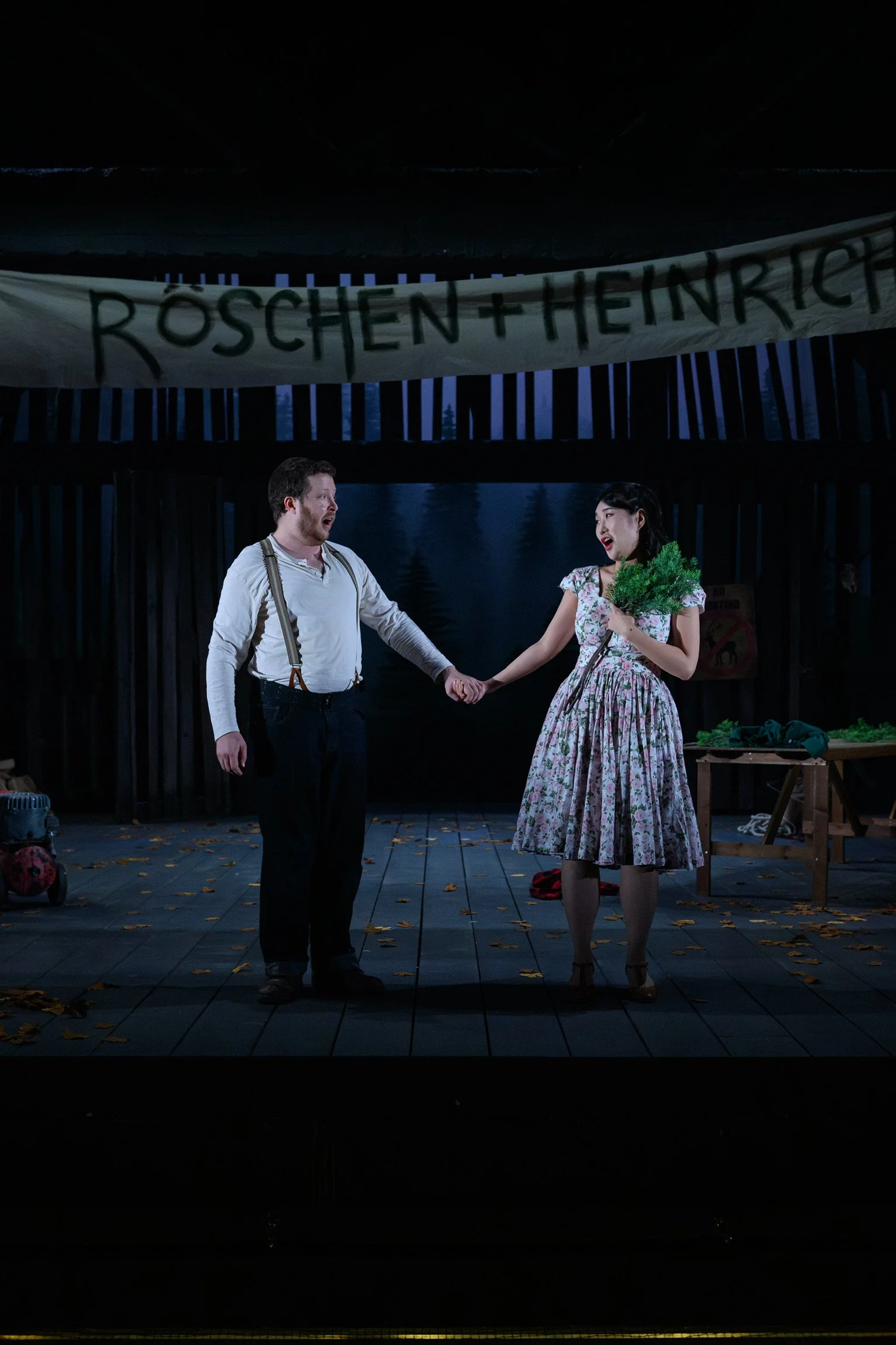 A man and woman holding hands on a stage, with a night background. The man is wearing suspenders and a white shirt, and the woman is holding a bunch of greens, wearing a floral dress. In the background, a banner reads 'ROSCHEN + HEINRICH'.