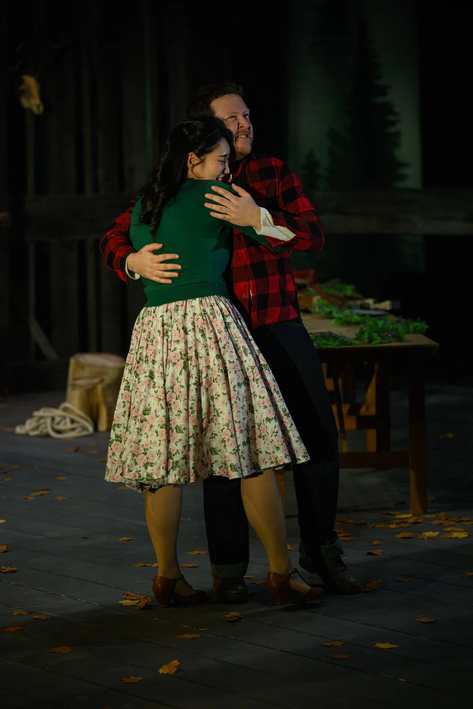 A man and woman hugging on a stage with fall leaves scattered on the floor, wooden furniture, and a dark forest backdrop.
