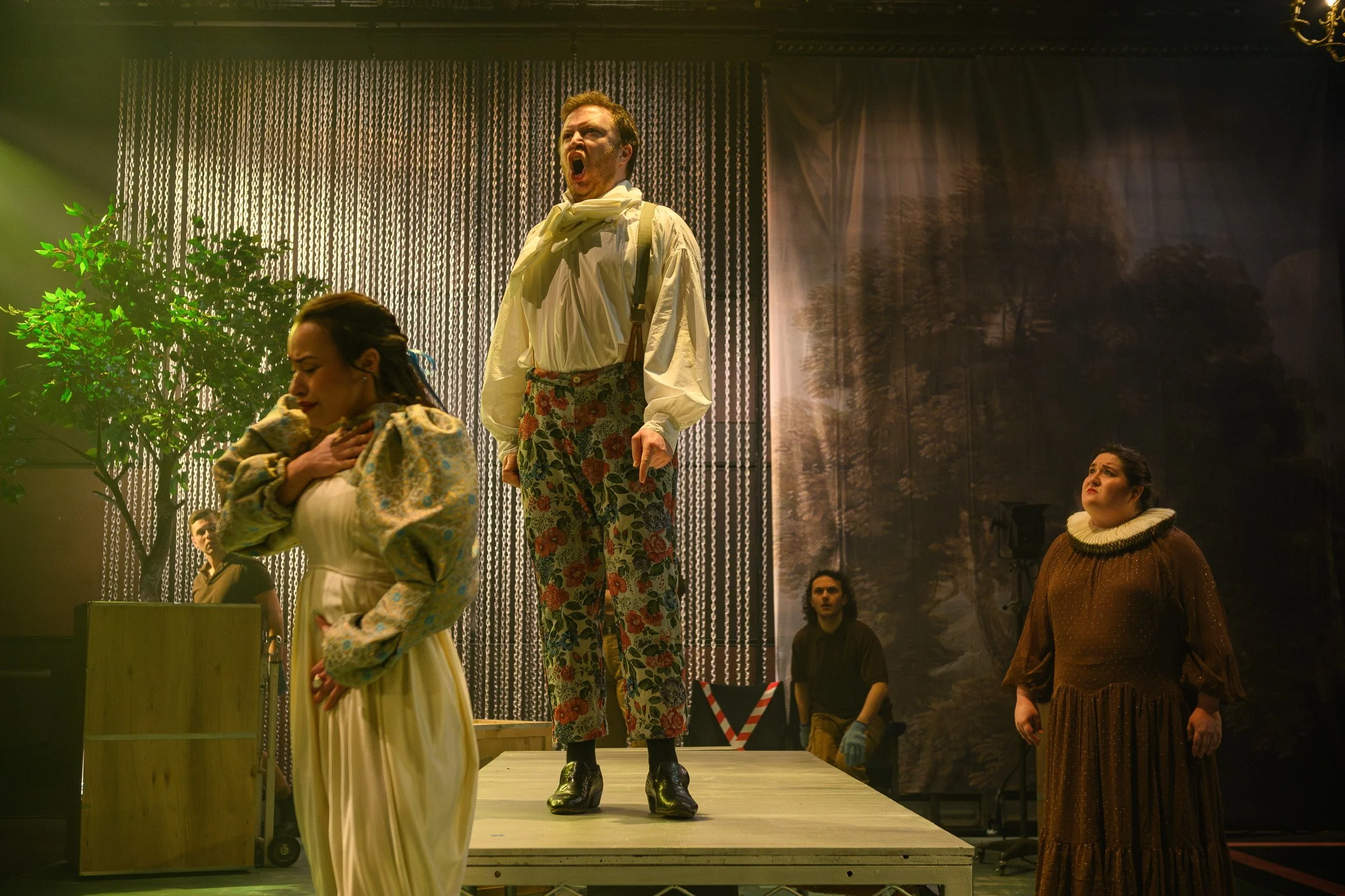 Actors performing on stage with dramatic expressions, dressed in elaborate costumes, in a theatrical setting with scenic backdrops and stage props.