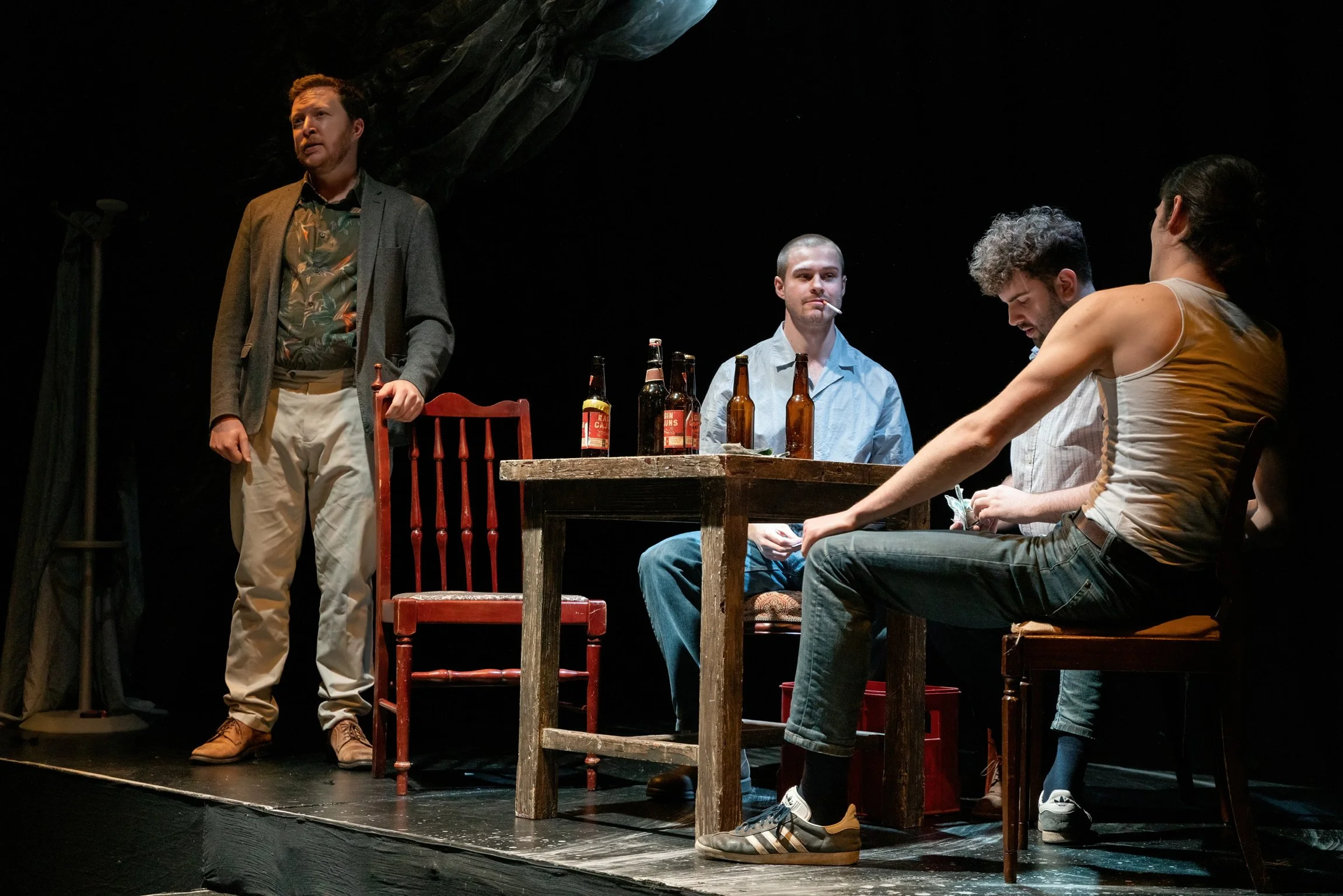 Four people are on a stage engaged in a theatrical performance. One man is standing while three others are sitting around a wooden table with bottles on it. The backdrop is black, and there is stage lighting.