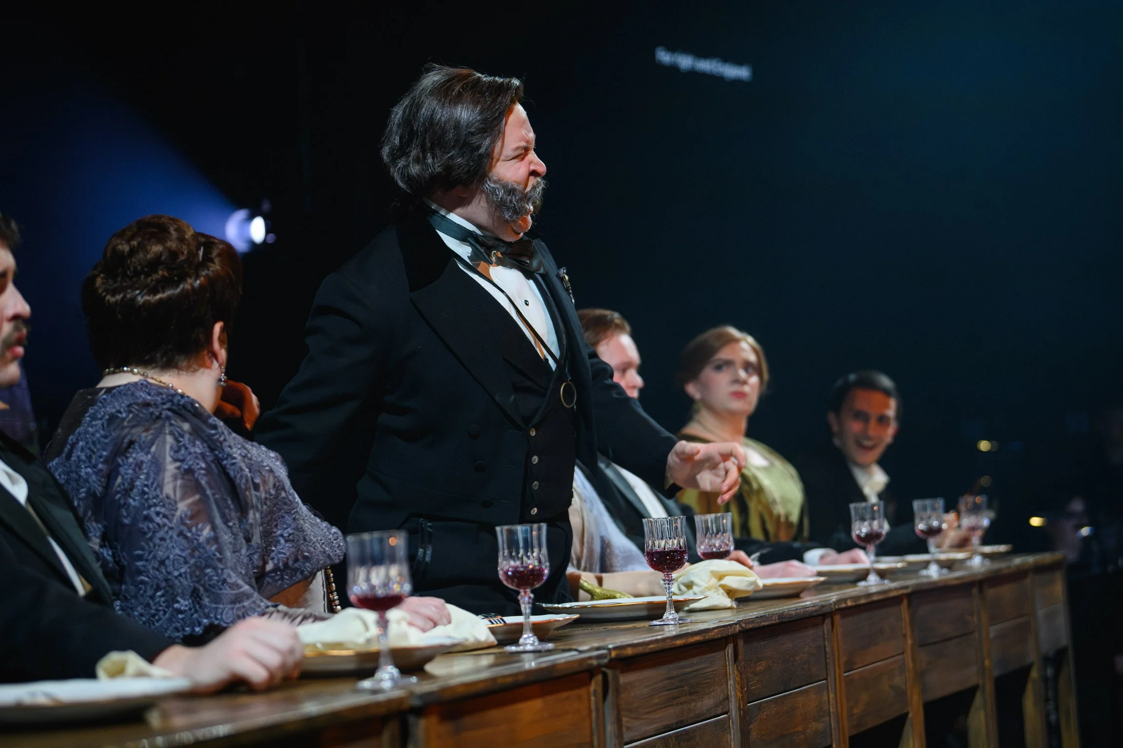 A man with gray hair and a beard, dressed in formal attire, is standing at a table during a formal dinner or event. He appears to be speaking or making a toast. Other seated individuals, mostly women, are visible around the table, with wine glasses i