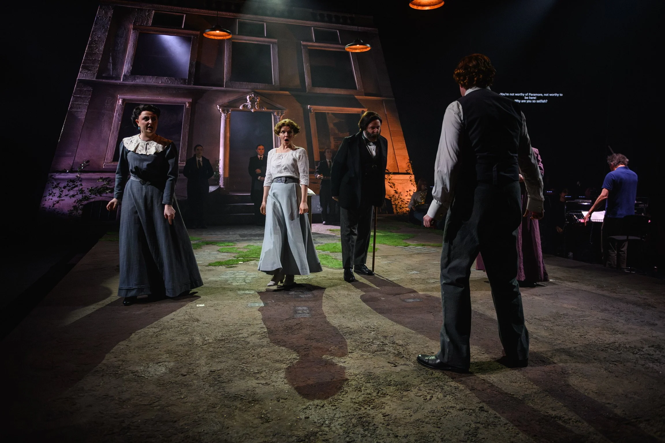 Actors performing on a stage, engaged in a theatrical scene with a backdrop of a house exterior, set in a dimly lit environment.