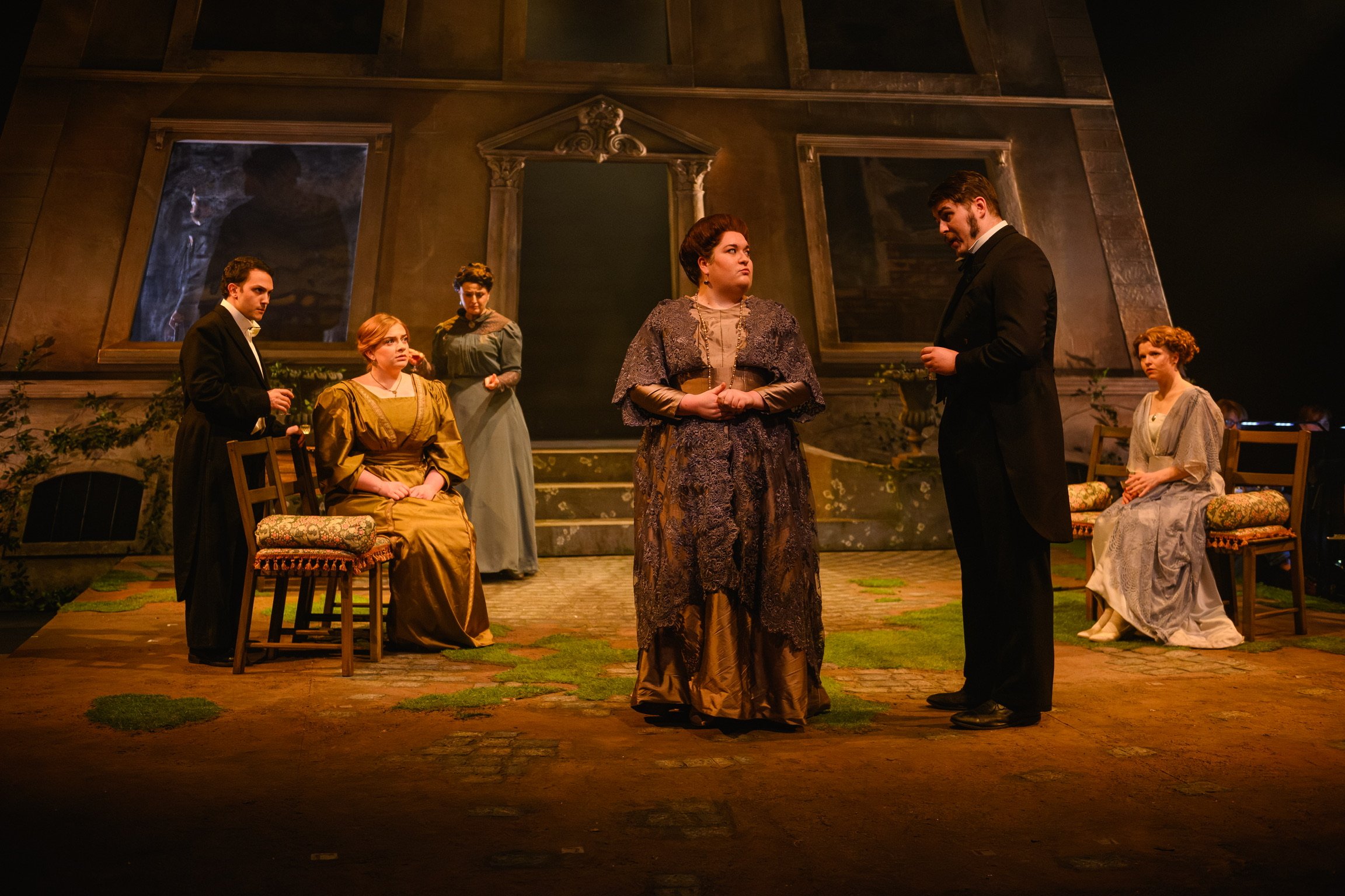 The scene depicts a theatrical performance set on a stage with six actors dressed in period costumes. The actors are positioned around a woman in a brown, ornate dress, who appears to be speaking or in a moment of emotional dialogue. The background f