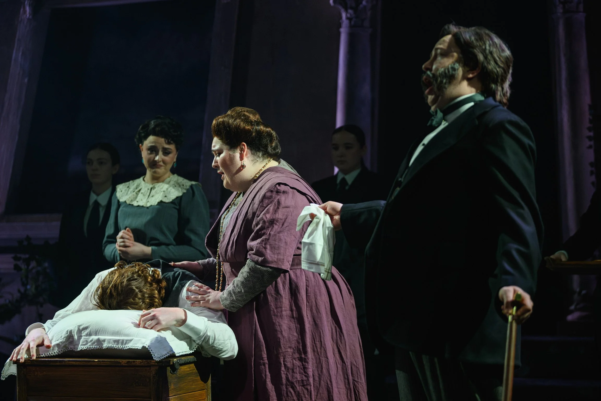 Scene from a theatrical performance featuring four women and one man on stage, with the woman in the foreground bending over a person laying on a bed or table, and the others standing behind her observing. The scene appears dramatic and historic, set