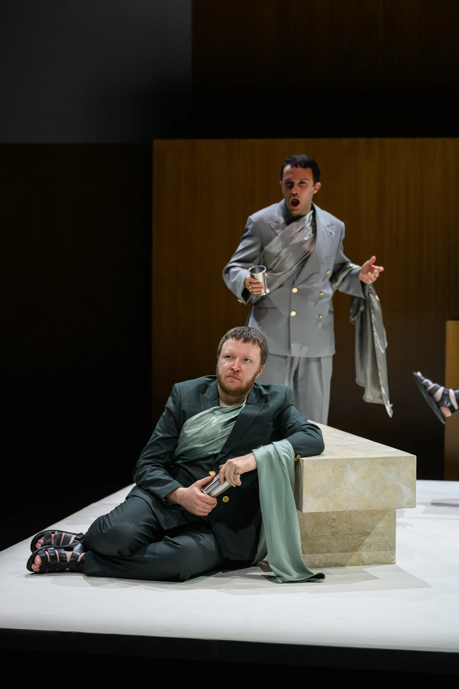 Two men dressed in suits are seen in a modern interior with one seated on the floor resting against a light-colored stone block with a draped fabric, and the other standing behind him, holding a glass and gesturing with his hand. Both men appear to b