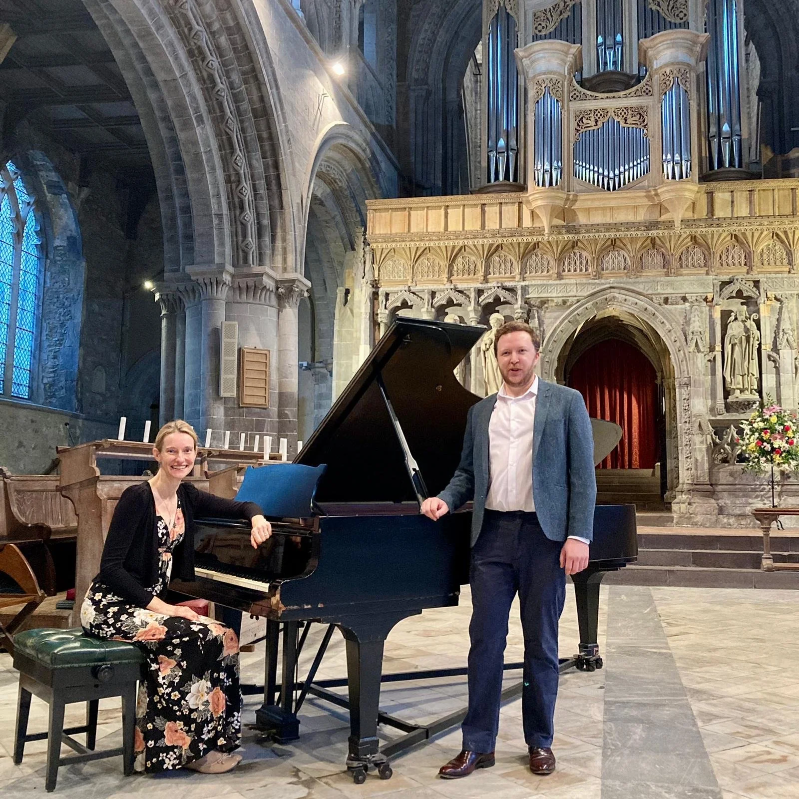 Two musicians, a woman playing the piano and a man standing beside it, inside a church with Gothic architecture, stained glass windows, and an organ in the background.
