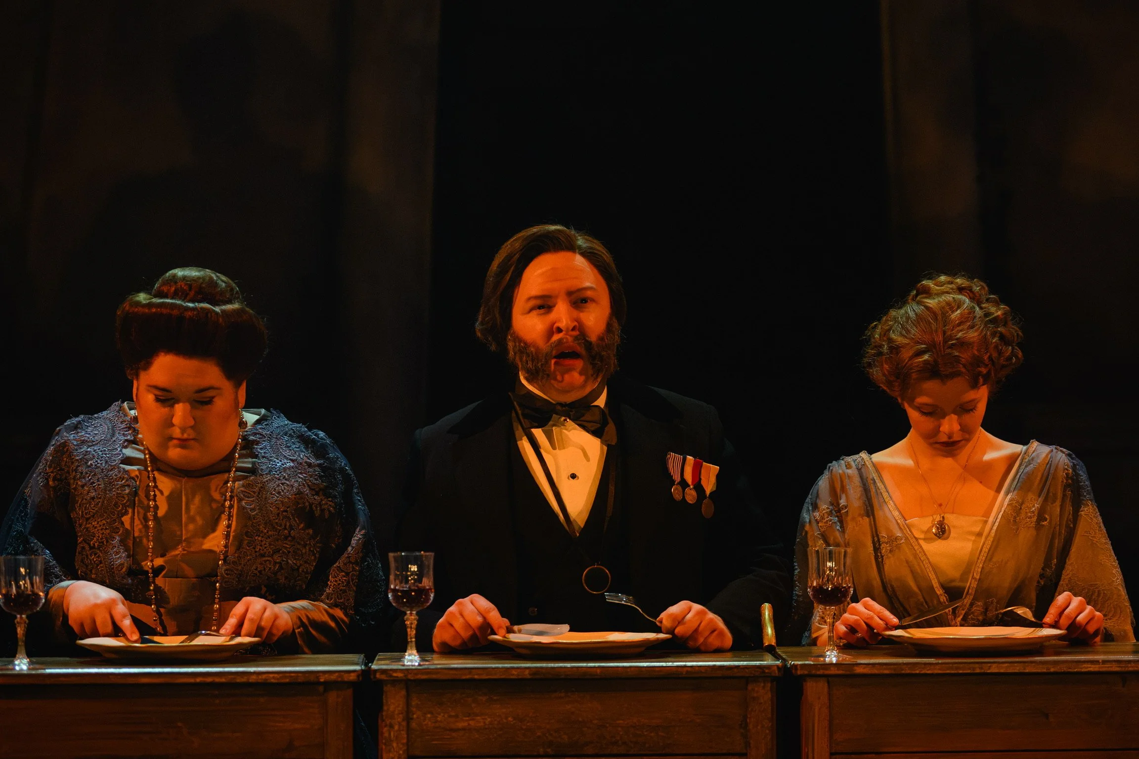 Historical scene with three people dressed in vintage clothing sitting at a dining table, one man in a tuxedo with medals, two women in period dresses, all bowing their heads in a somber moment.