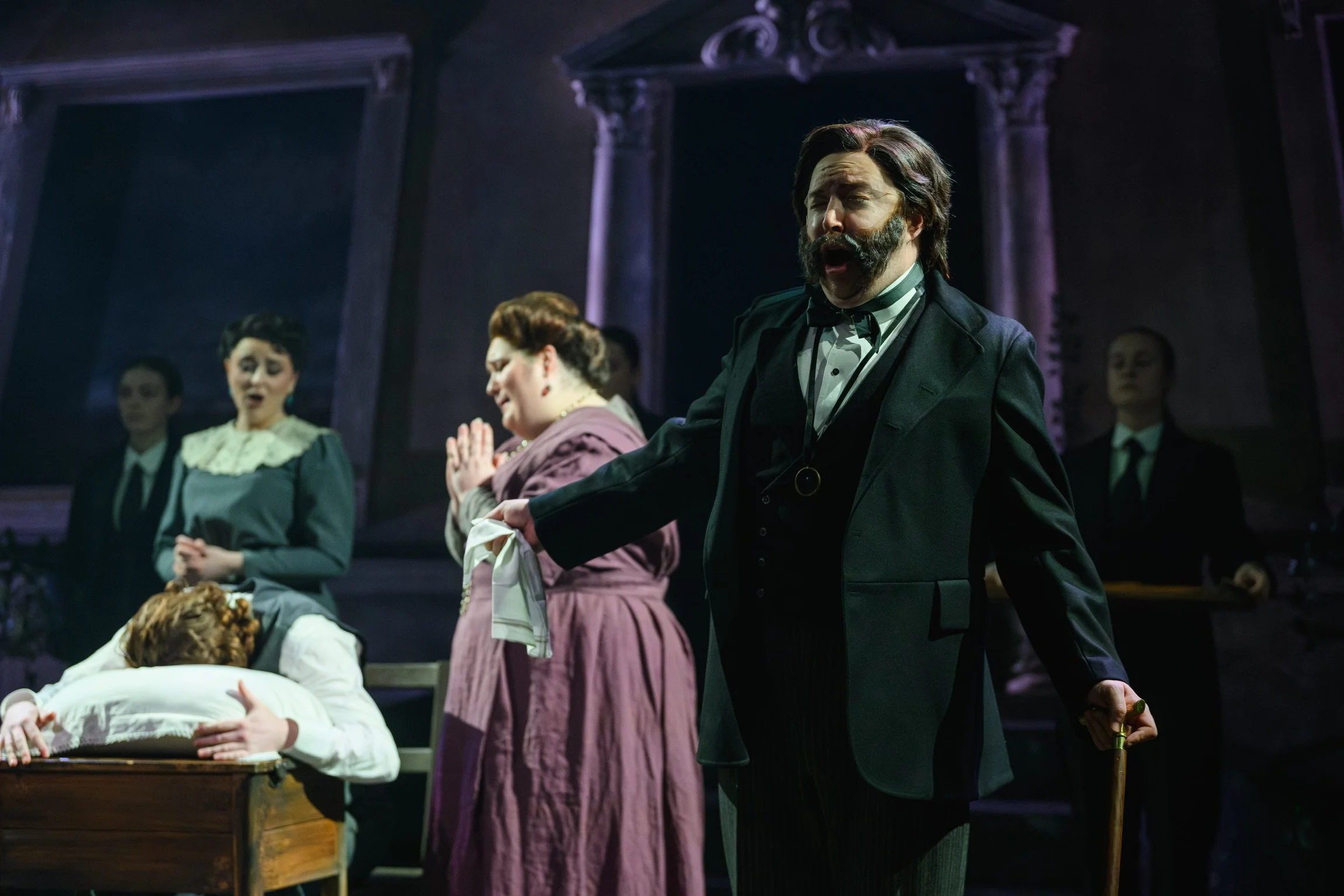 A theatrical scene with actors dressed in period costumes on stage, including a man with a beard and mustache singing or speaking with others in the background.