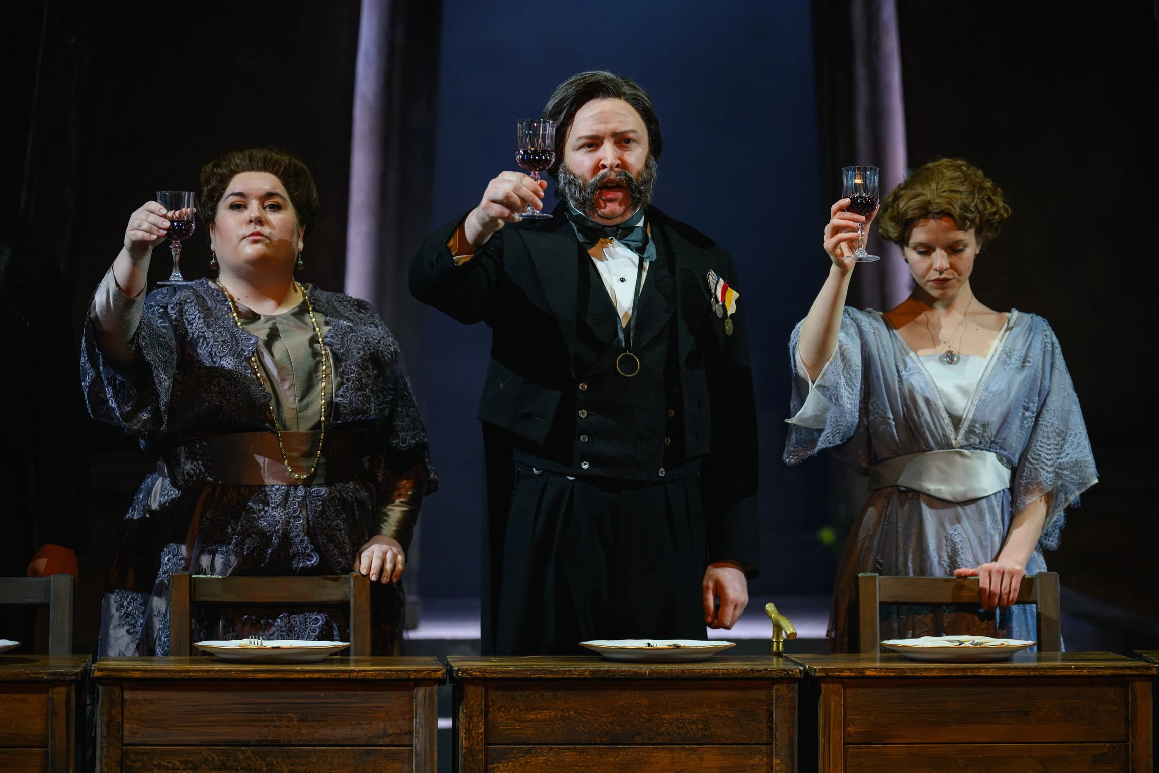 Three actors in period costumes stand behind a wooden dining table, raising glasses of red wine in a theatrical scene. The man in the center has a beard, mustache, and is dressed in a black tuxedo with medals. The woman on the left has dark hair styl