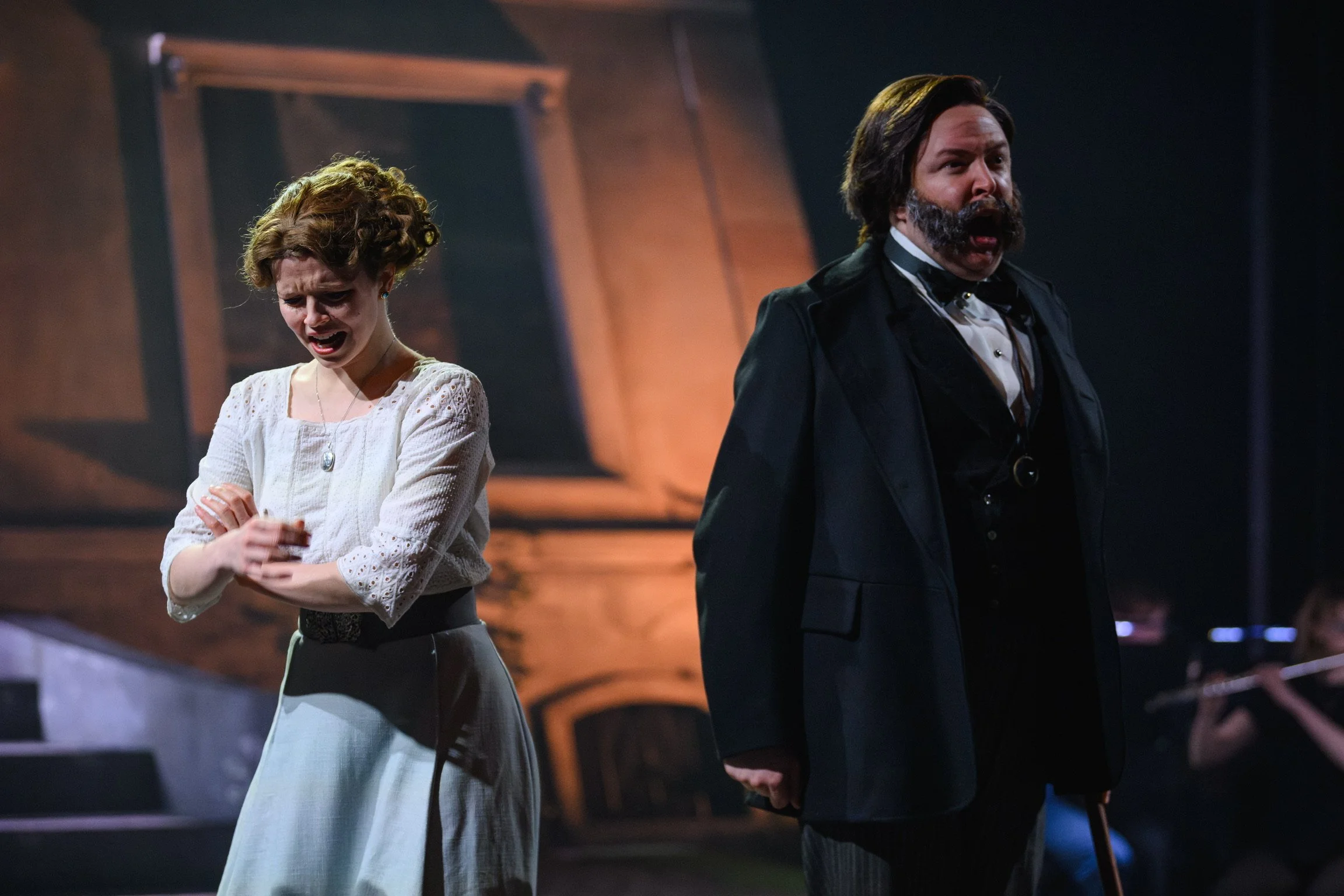 Two actors on stage, one woman with curly hair and a white blouse looking distressed, and one man with dark hair and a beard in a black suit with a bow tie shouting or singing, holding a cane. The background appears to be a theater set.
