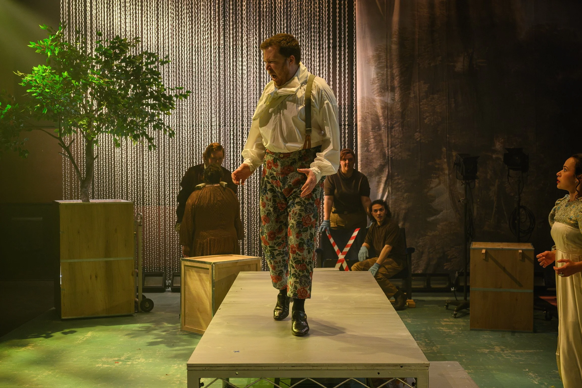 A man with a beard and mustache, wearing floral pants, a white shirt, suspenders, and black boots, is shouting or singing on a stage with several people in the background. There is a tree on stage, and the backdrop is decorated with metallic chains a