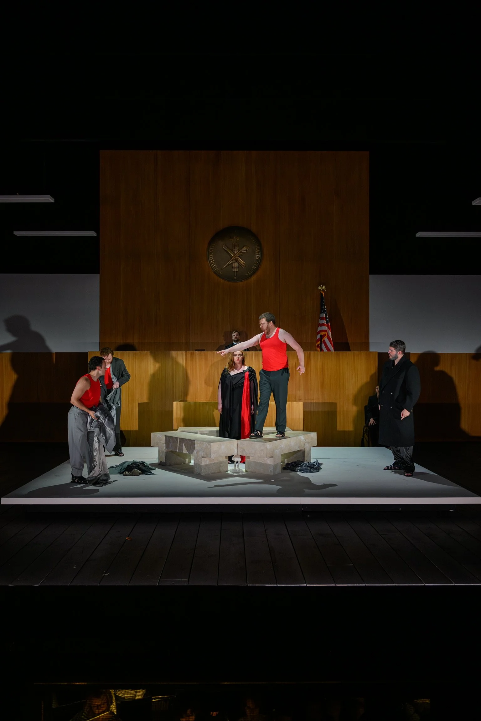 The image shows a theatrical performance or a play with several actors on stage. The set features a raised platform or altar with some clothing items on the floor, and the background includes a large wooden panel, a clock, and an American flag. The a