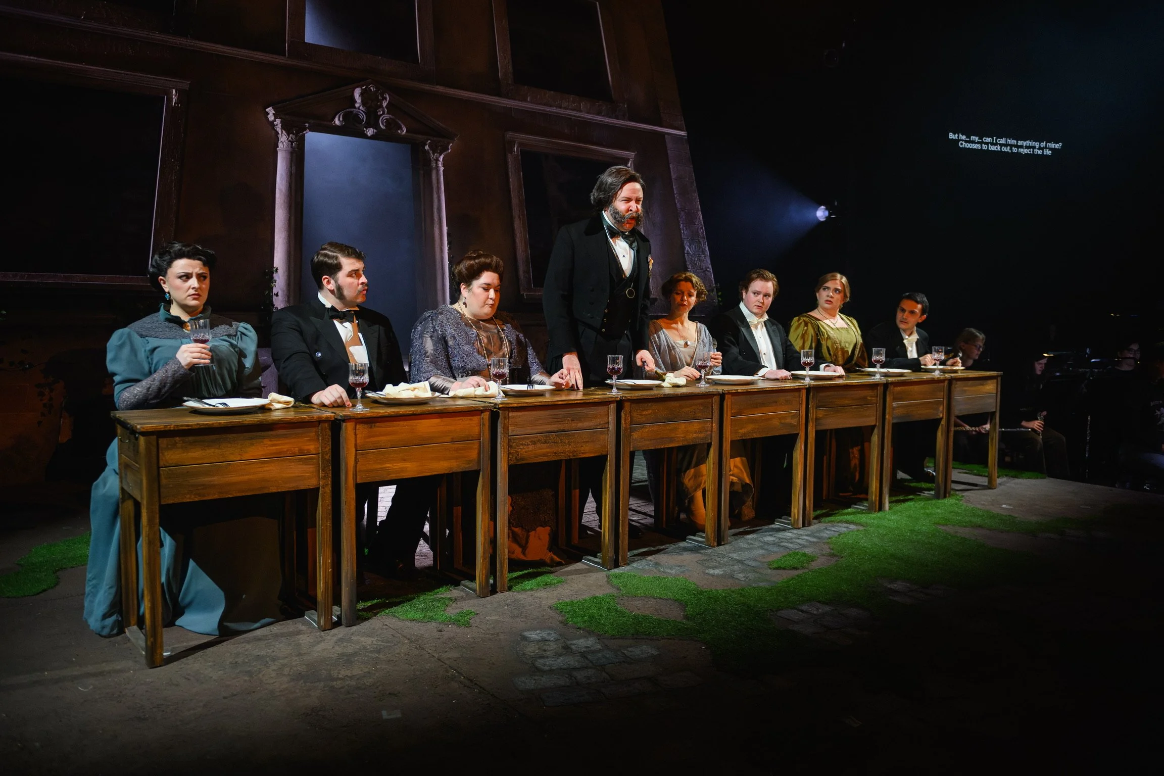 A theatrical performance with seven actors in period costumes sitting at a long wooden table, with one standing man speaking passionately, set against a dark stage with a backdrop of large wooden frames.