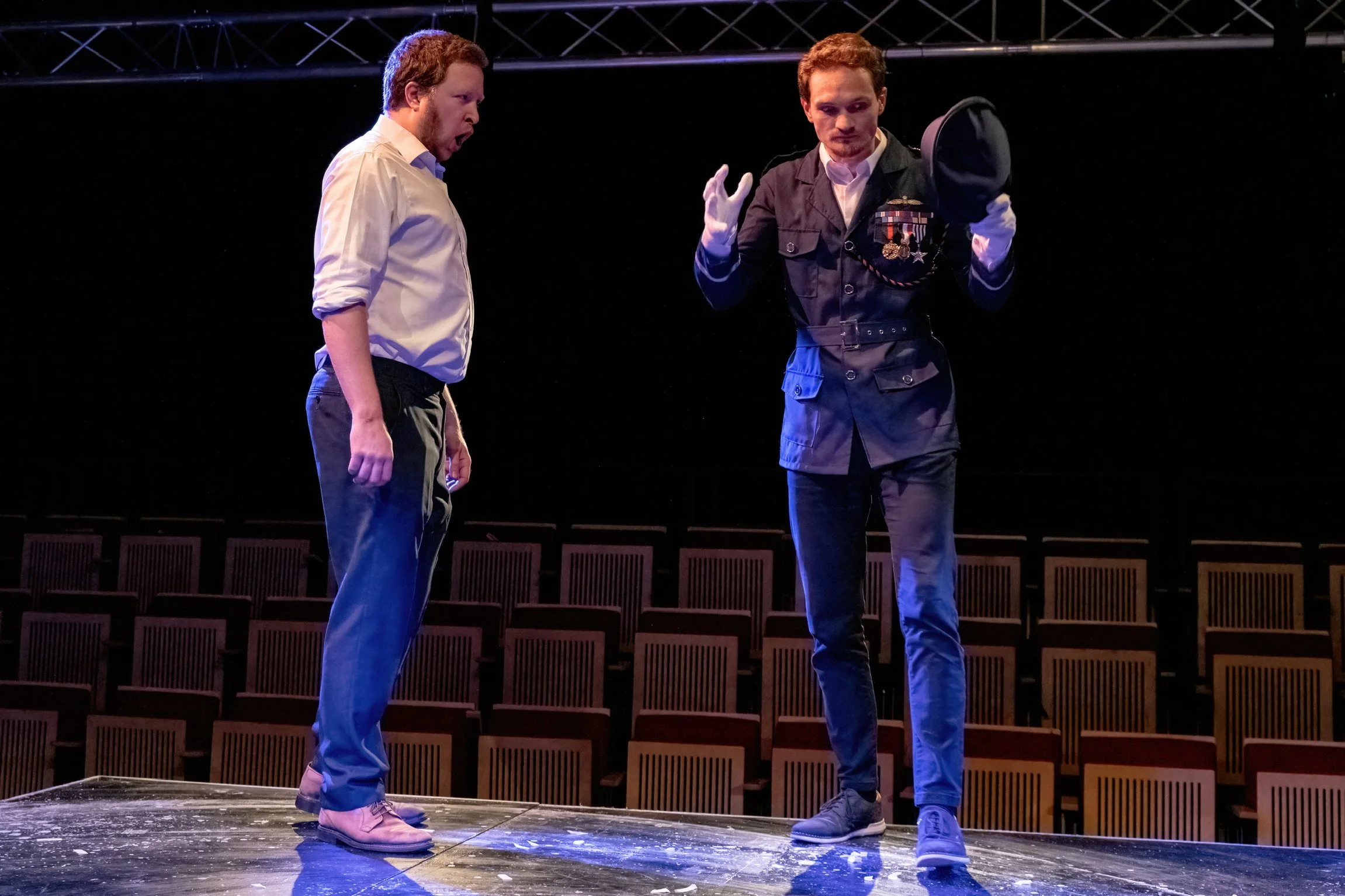 Two men standing on a stage, one in a white shirt and the other in a pilot uniform, engaged in a theatrical performance or rehearsal.