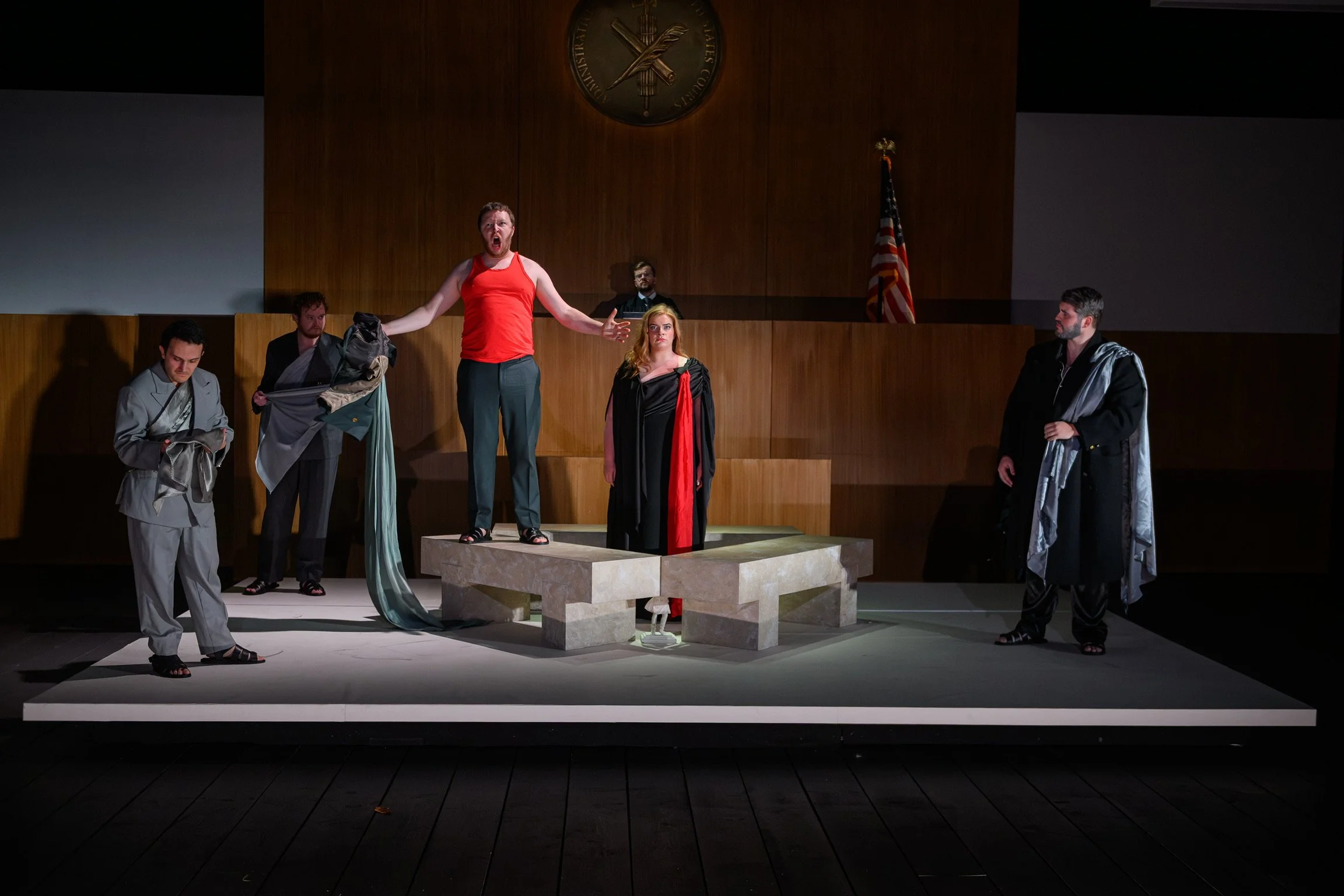The image shows a theatrical stage scene with five actors in costume. One actor in a red sleeveless top is standing on a platform, appearing to shout or speak passionately, with arms outstretched. The others stand around, some holding clothing or pro