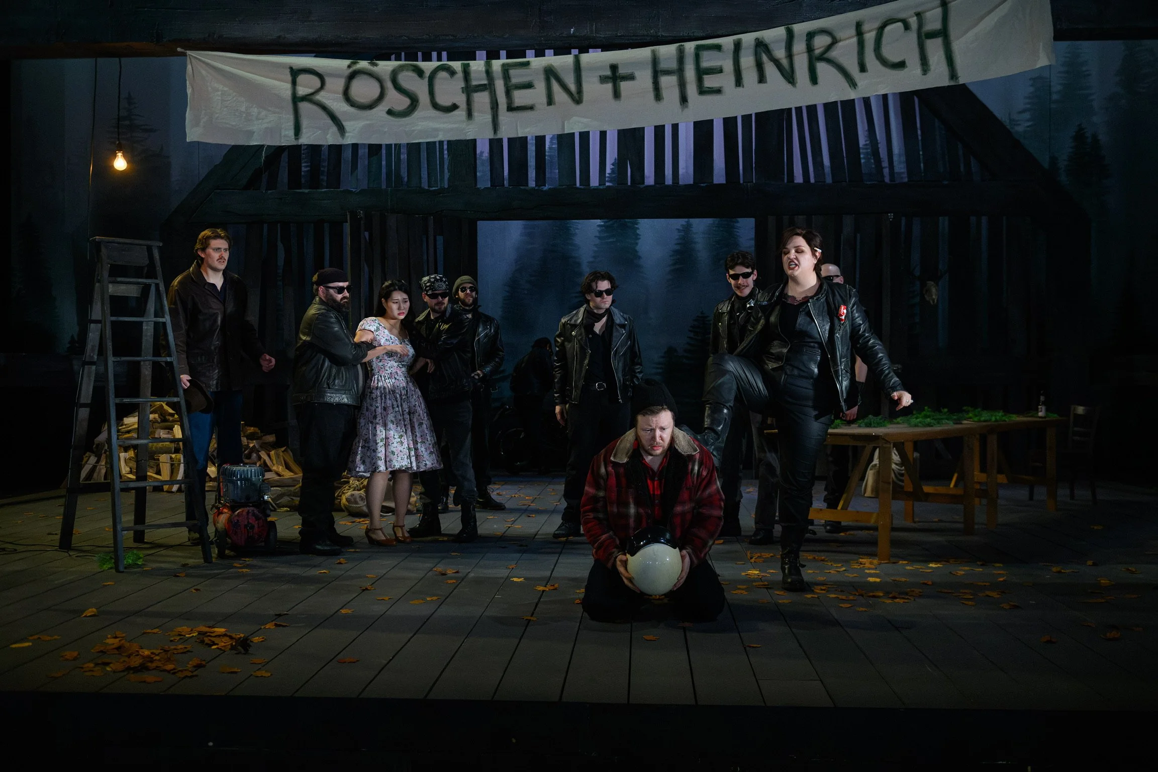 A theatrical stage scene with several actors dressed in leather jackets, some wearing sunglasses. One woman is making a fierce expression, and a man in the foreground is sitting on the floor holding a white helmet. The background features a forest sc