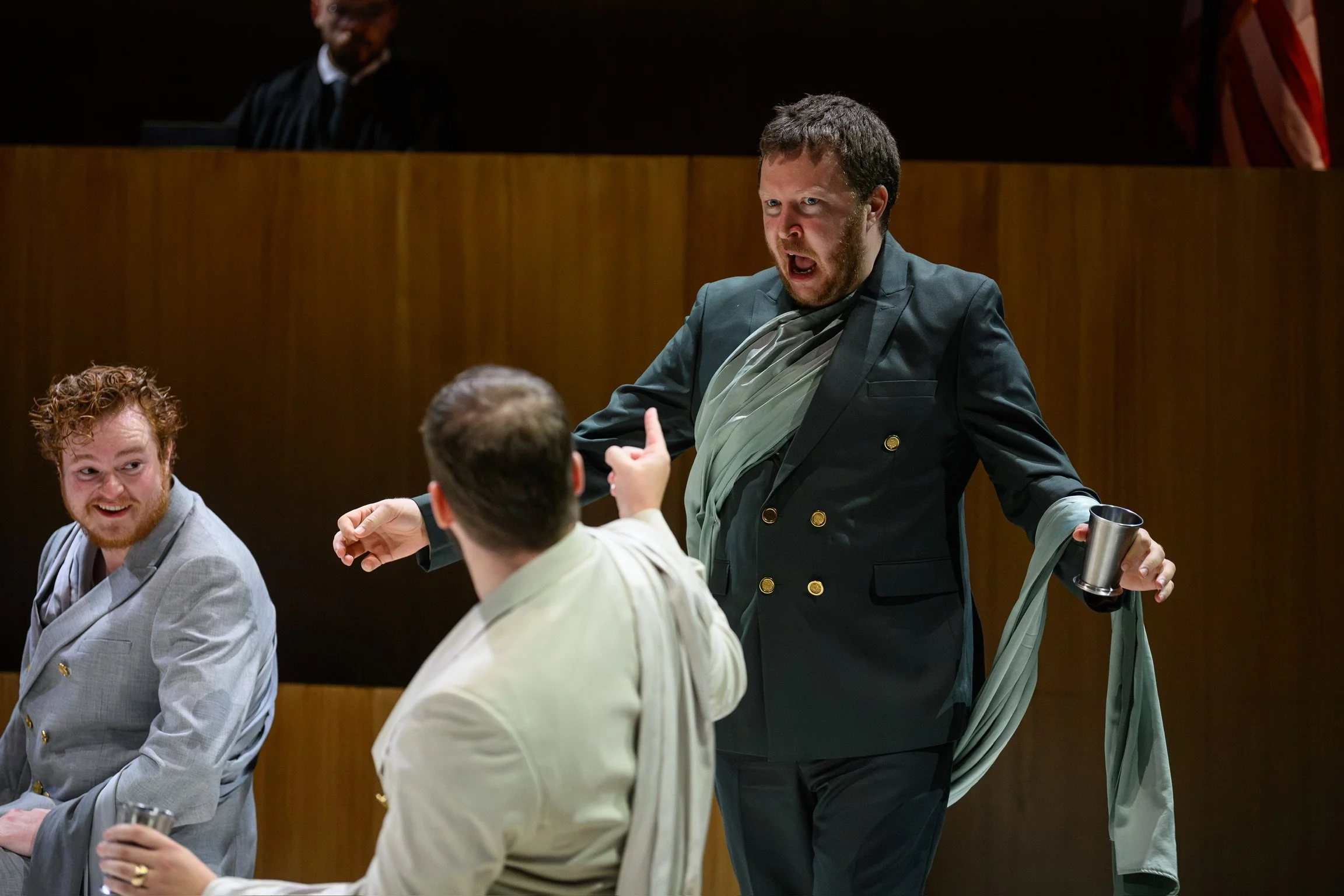 A man with a beard and short hair, wearing a dark suit with gold buttons, appears to be shouting or arguing with a person with short hair, dressed in a light-colored shirt. The man in the suit is holding a metal tumbler. There is also another man wit