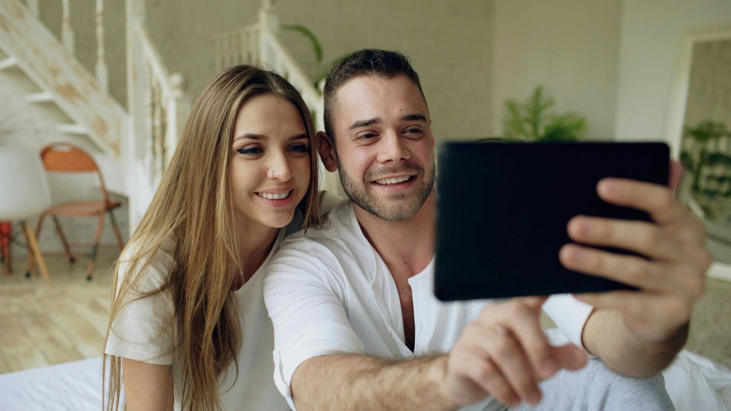 A young couple taking a selfie together in a bright, cozy room with a staircase and chairs in the background.