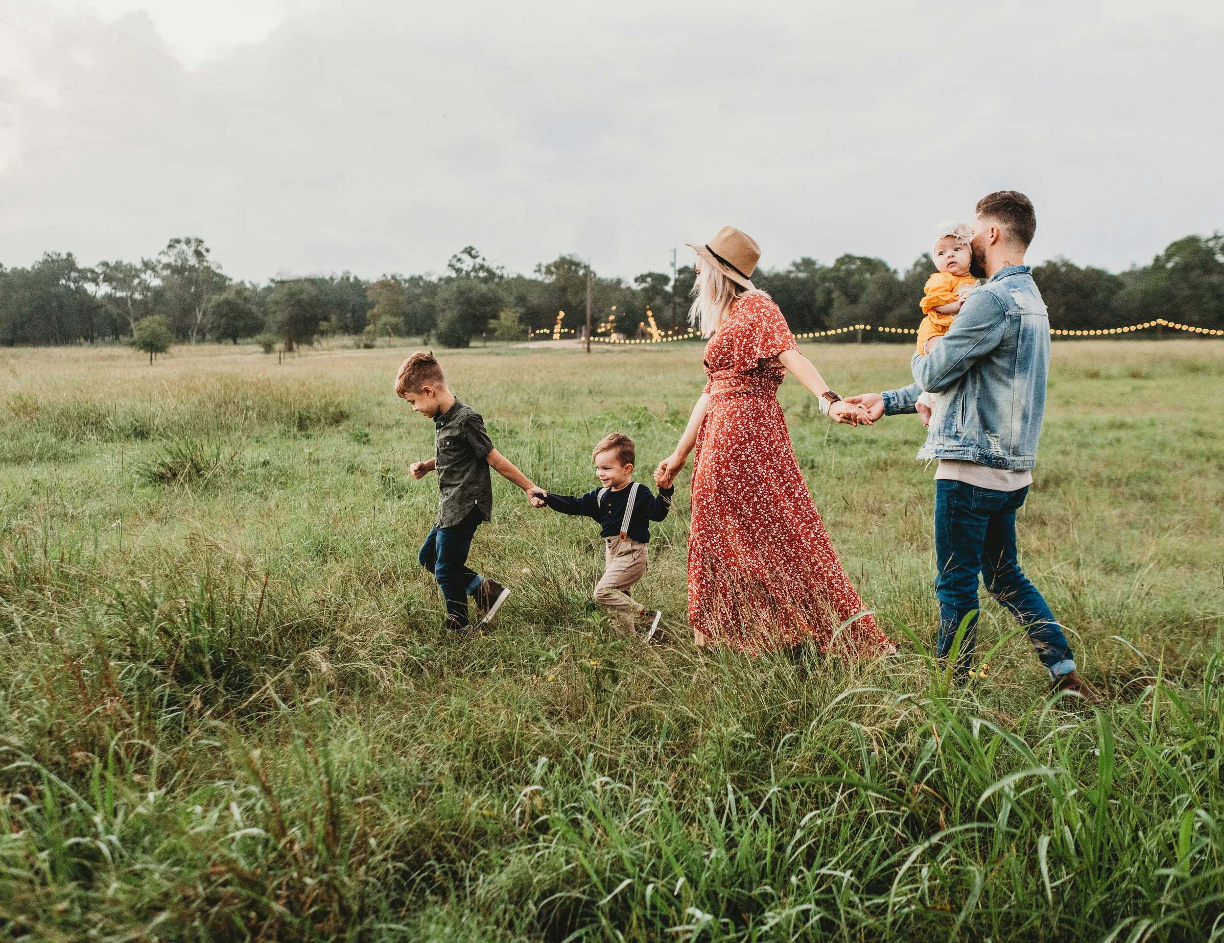 A group of children and a man holding a baby walk through a grassy field with trees in the background and string lights hanging in the distance, under a cloudy sky.