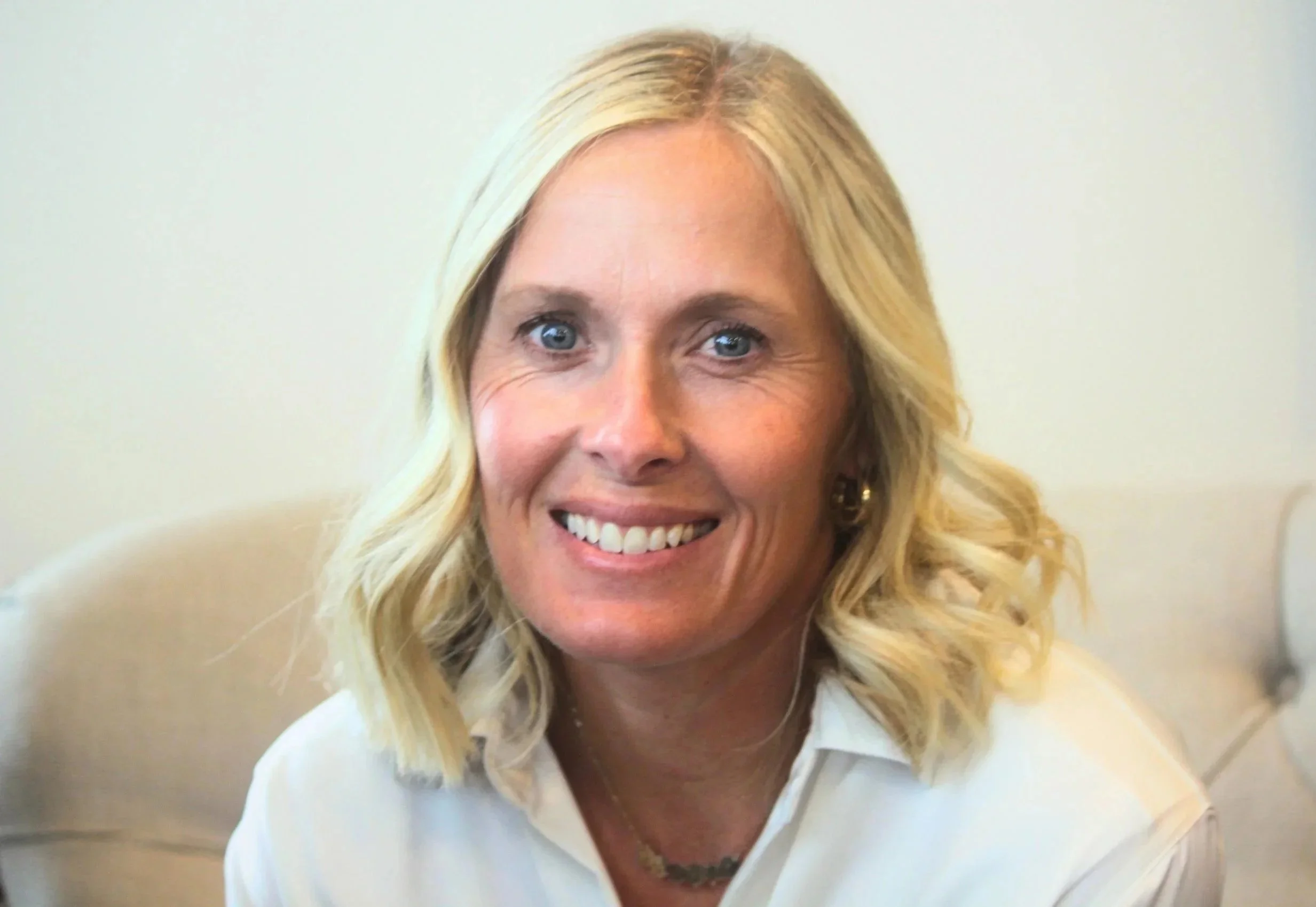 Close-up of a smiling woman with blonde, wavy hair, wearing a white blouse, against a plain light-colored background.