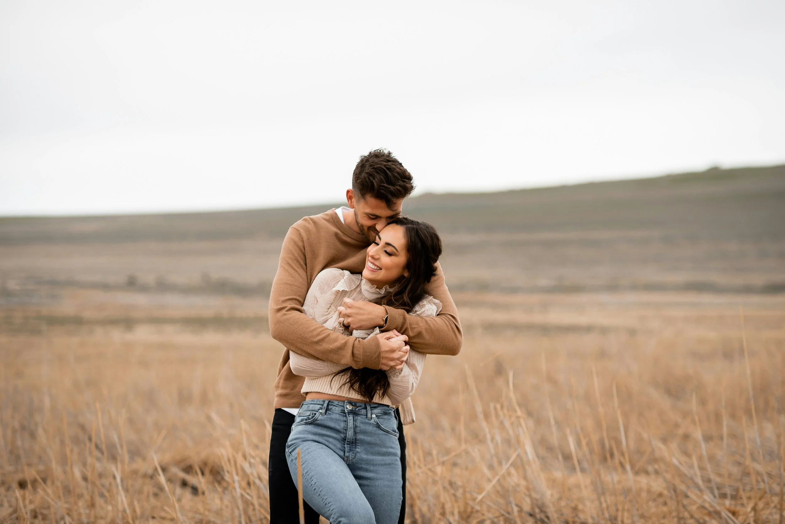 A couple embracing and smiling in a field of tall grass with a cloudy sky.
