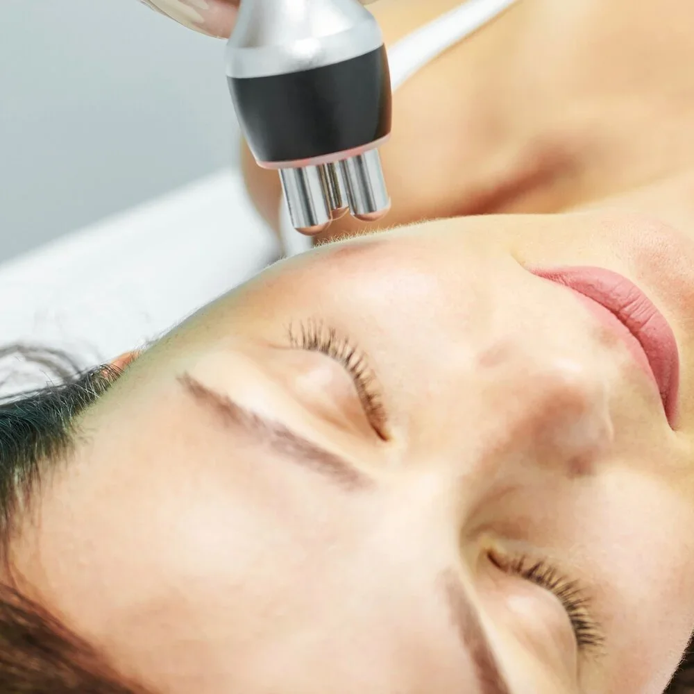 A person receiving a facial treatment with a device held above their face while lying down with eyes closed.