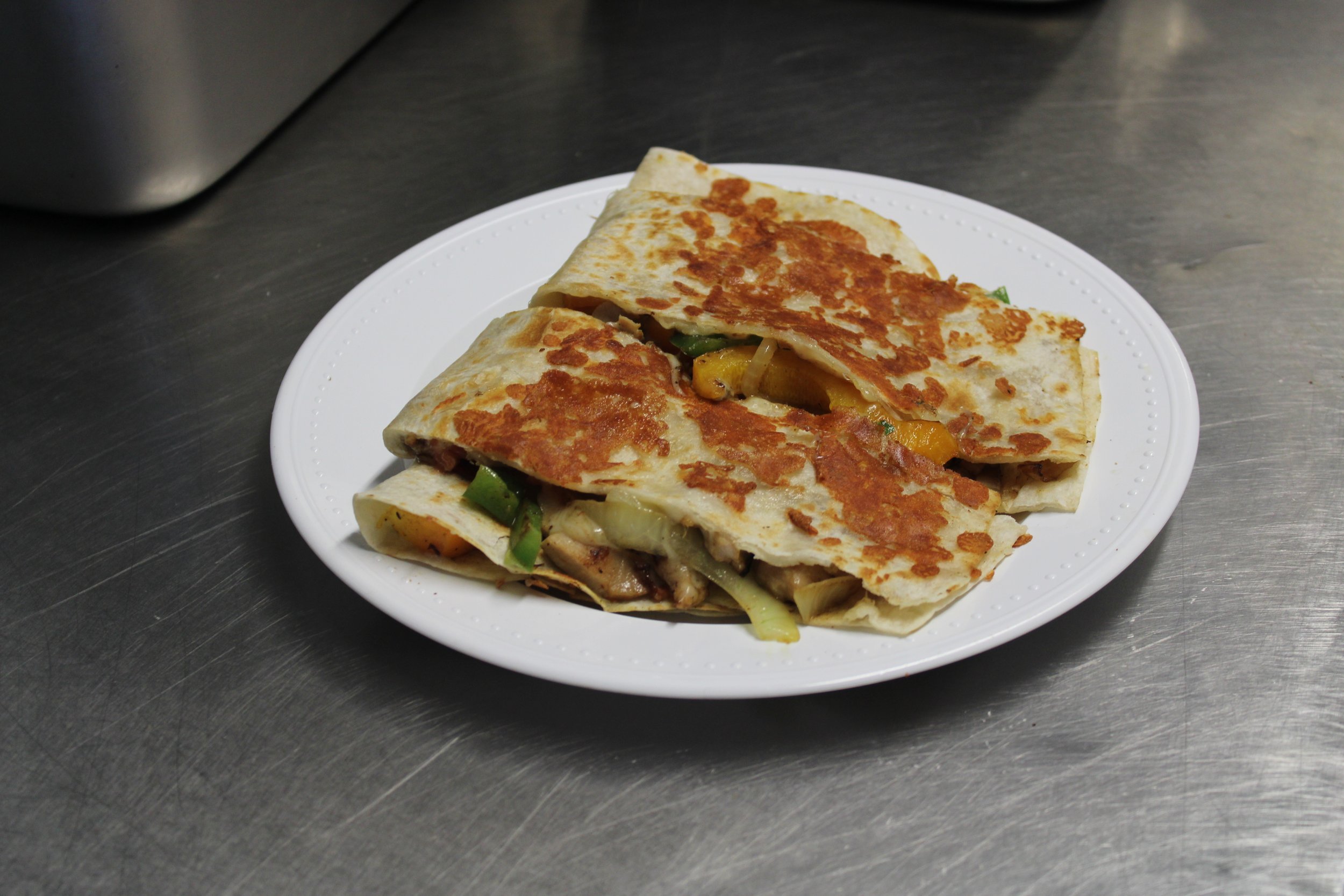 Quesadilla on a white paper plate, with melted cheese and vegetables inside, on a stainless steel surface.