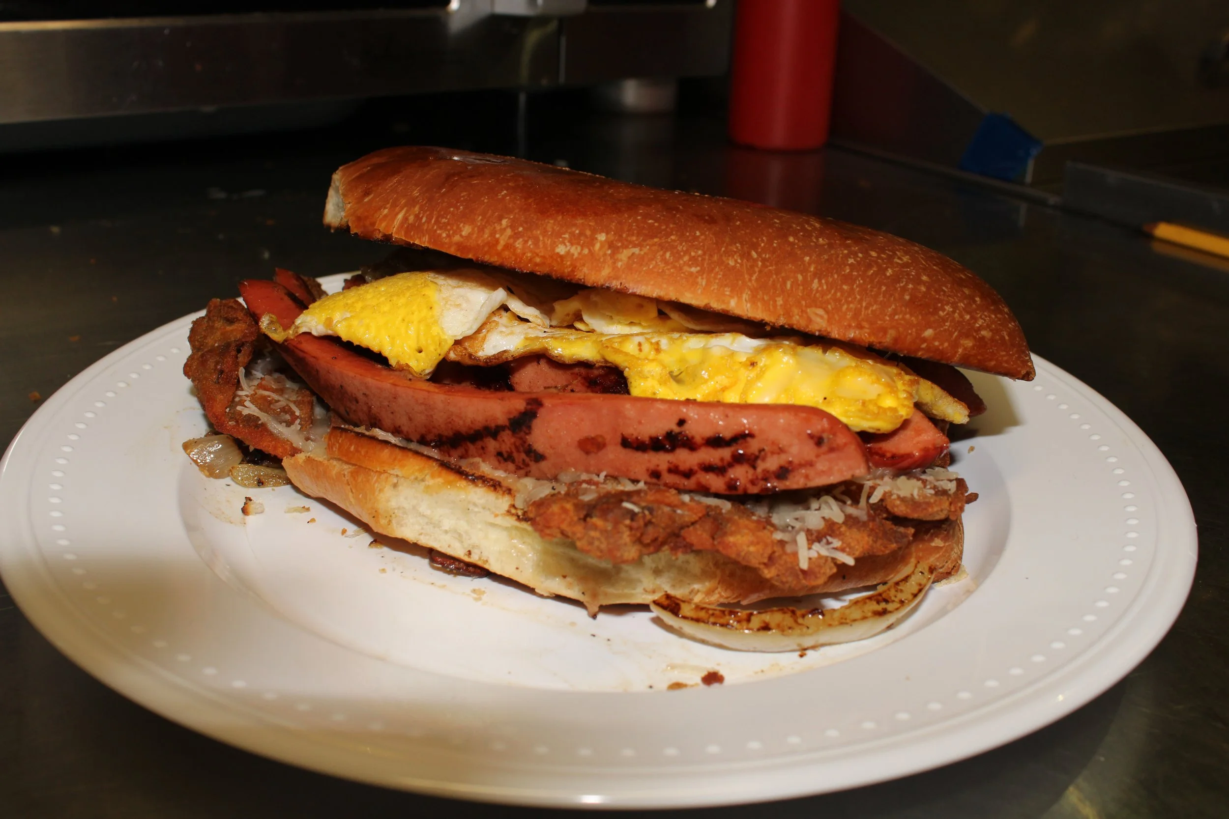 A sandwich with fried chicken, fried egg, slices of ham, bacon, and onions on a hoagie bun on a white plate.