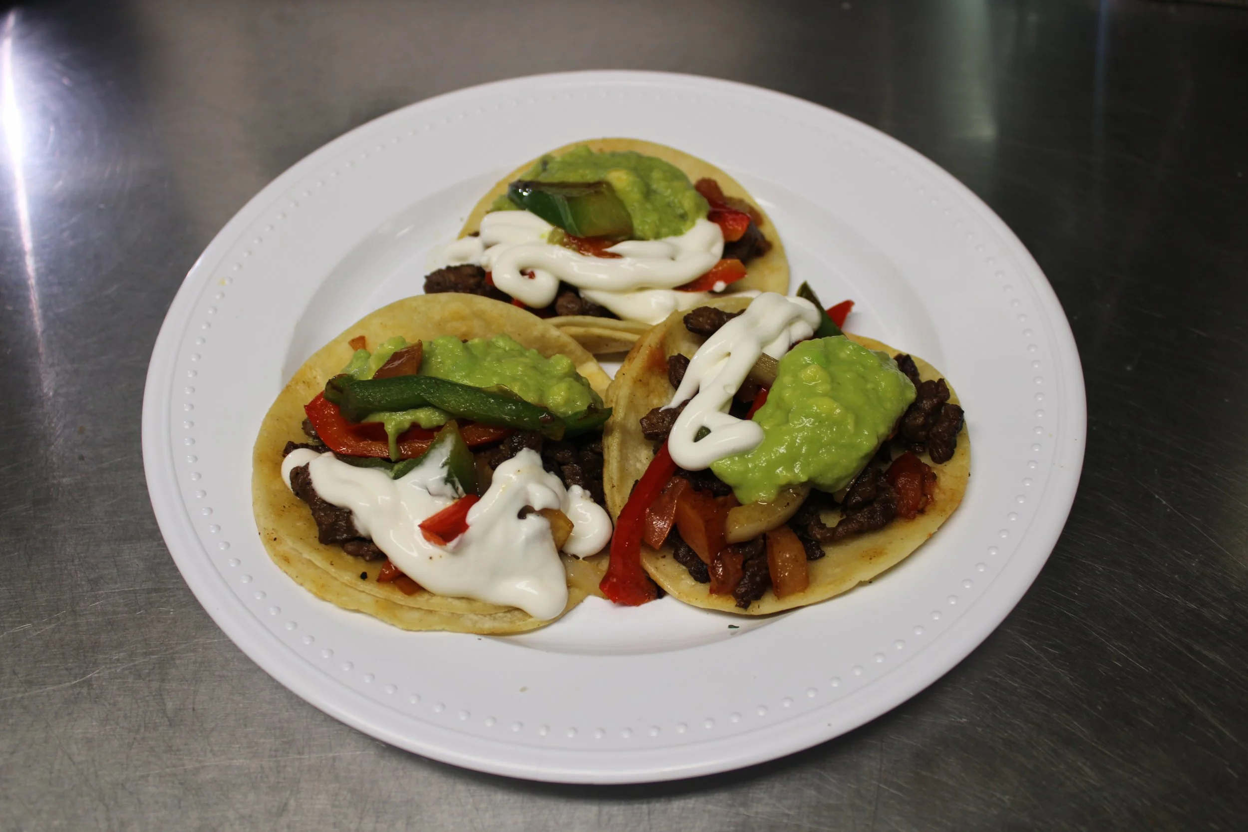 Three street tacos with beef, diced red and green peppers, guacamole, and mayonnaise on a white plate.