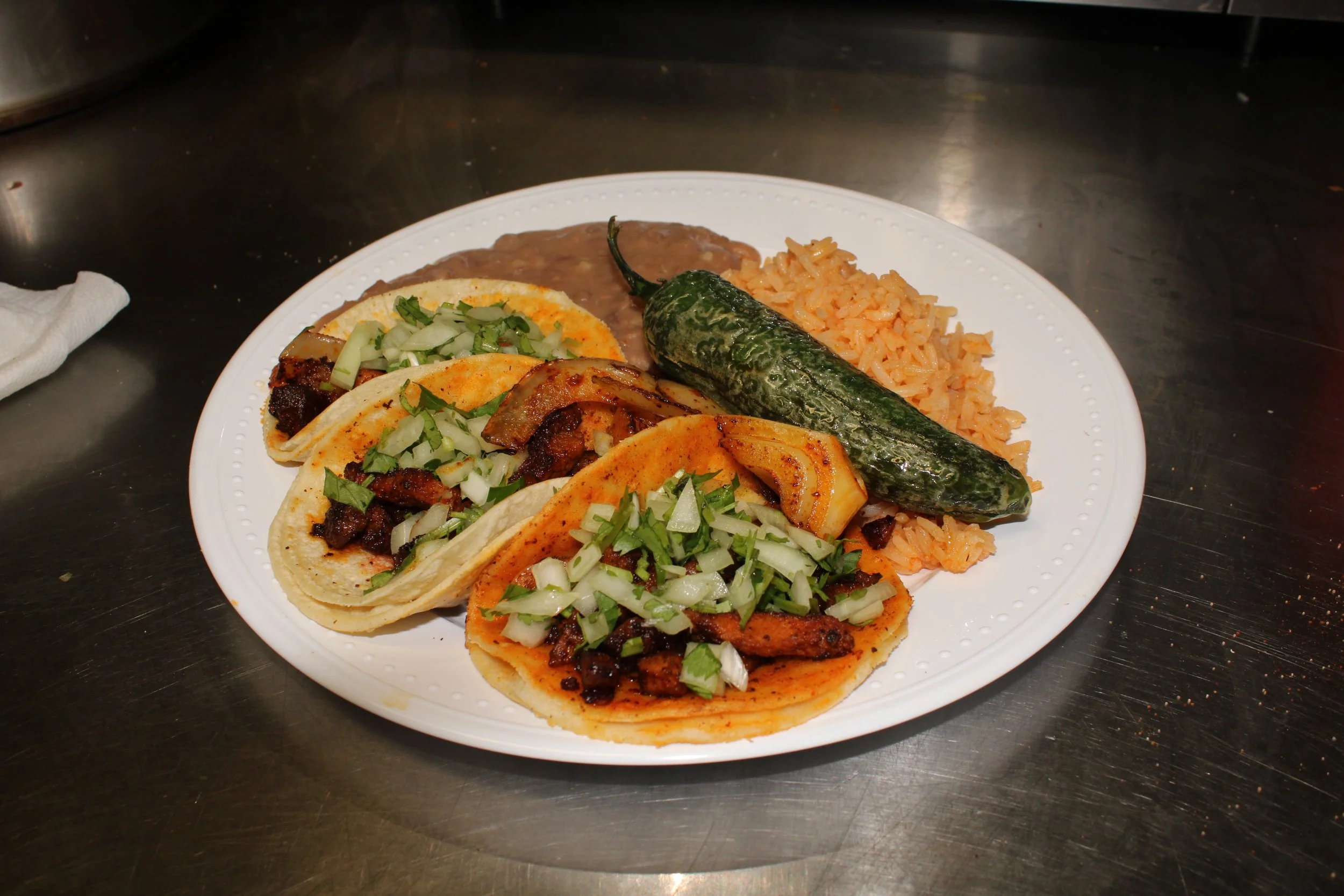 Plate with three tacos filled with meat, chopped onions, and cilantro, a grilled jalapeño pepper, and rice, placed on a dark surface.