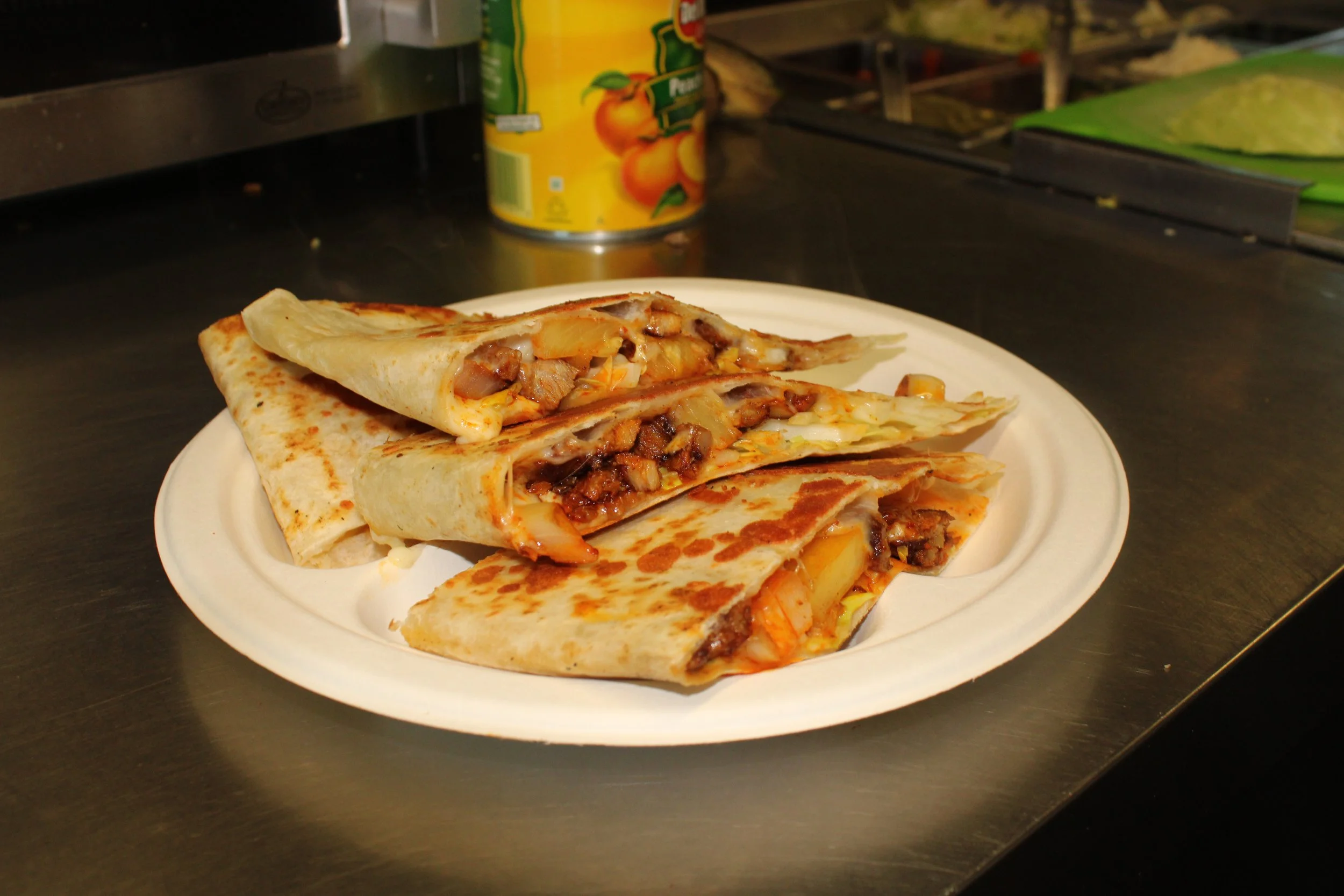 A plate of two cuts of Mexican food, possibly burritos or quesadillas, filled with cheese, meat, and vegetables, placed on a white paper plate. In the background, there is a canned fruit juice and a green container on a dark countertop.