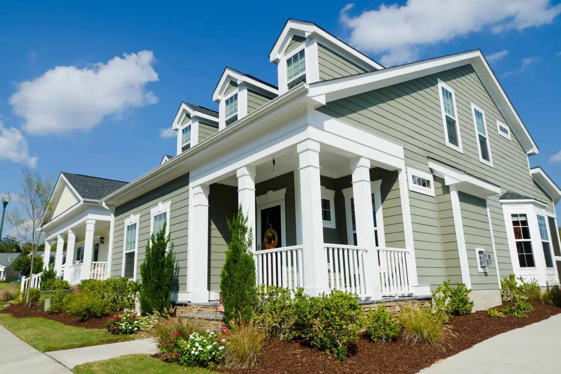 A two-story house with green siding and white trim, featuring a covered porch with white columns, surrounded by landscaped garden with shrubs, flowers, and a sidewalk under a partly cloudy blue sky.