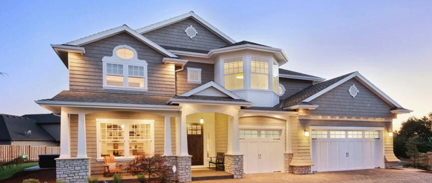Large two-story house with beige siding and stone accents, white trim, covered front porch, and three-car garage, illuminated at dusk.