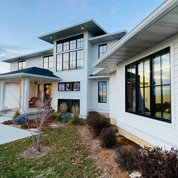 Modern multi-story house with large windows, white exterior, and a landscaped yard with shrubs and a small tree
